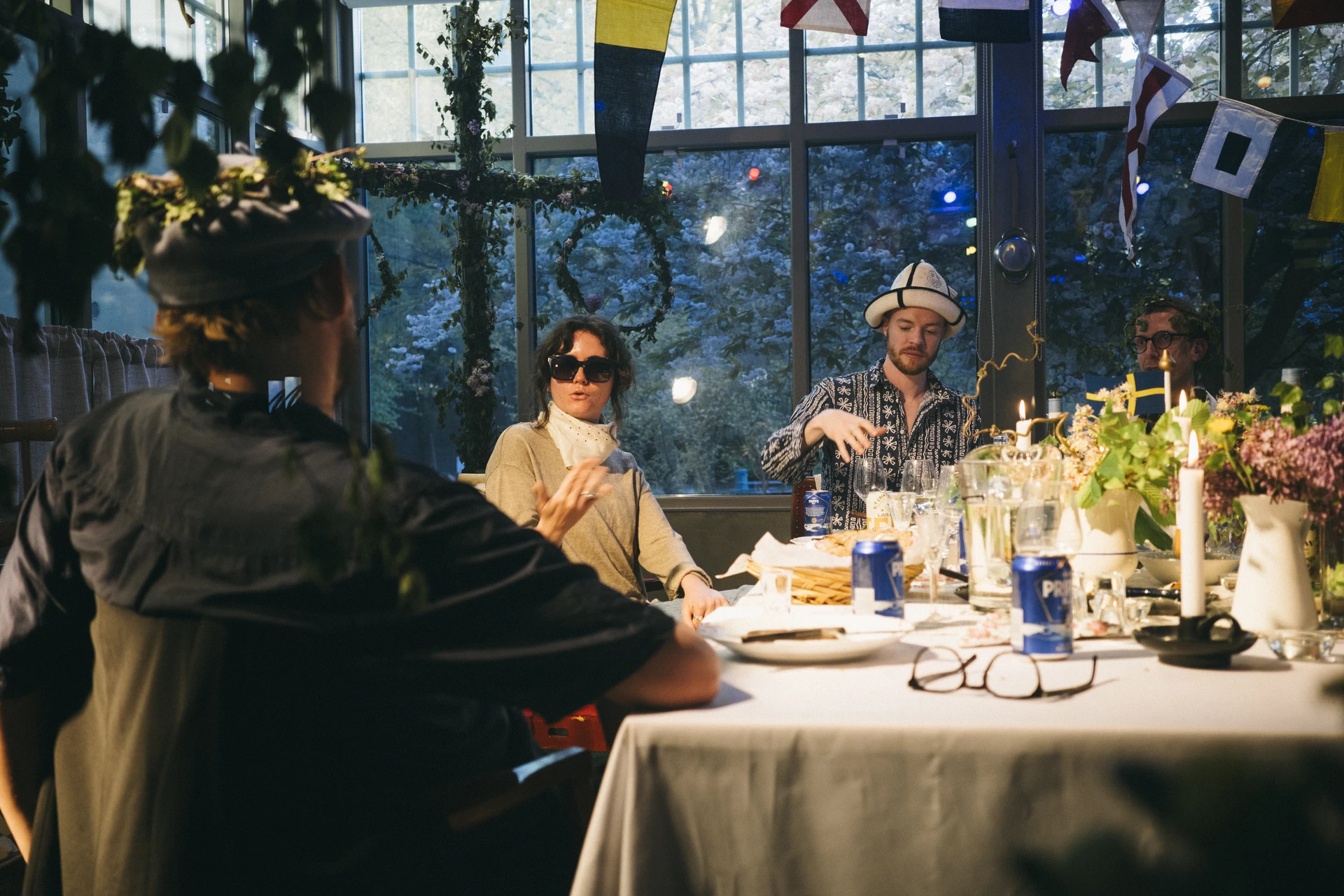 Guests in hats and flowers sit at a long candlelit table under greenery and bunting, in front of tall windows.