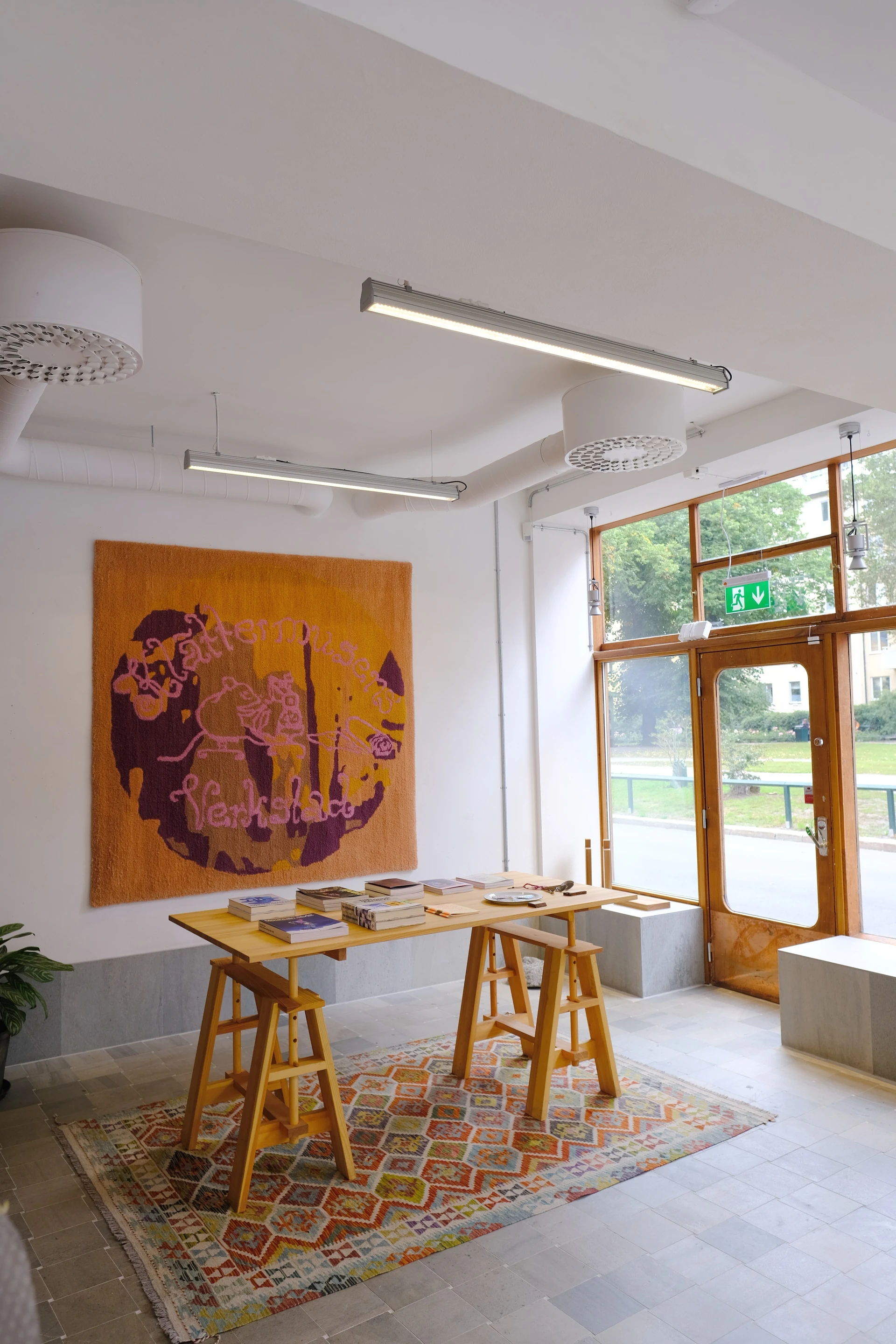 The finished rug hangs above a table of books in Klättermusen's Verkstad, with the park visible through the open door.