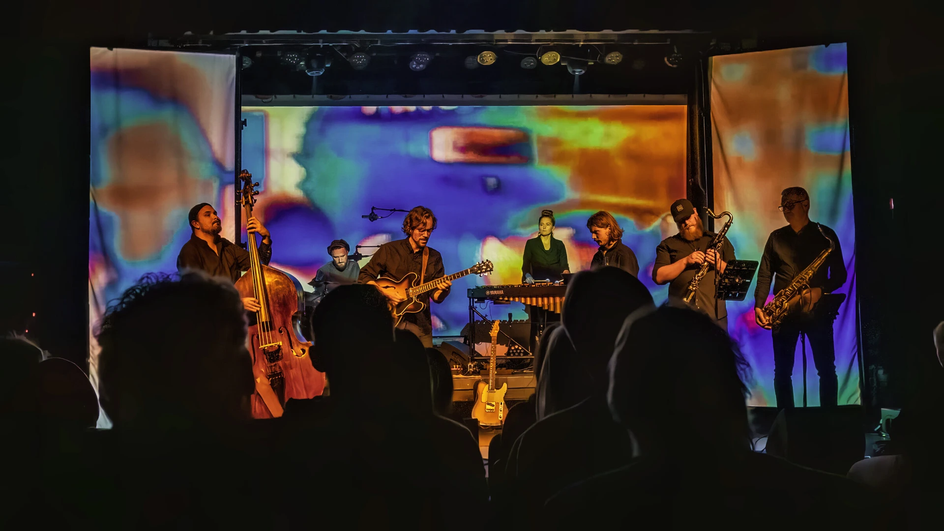 A seven-piece jazz band plays in front of three hanging screens washed in blurred purple, orange and turquoise projections, the audience in silhouette.