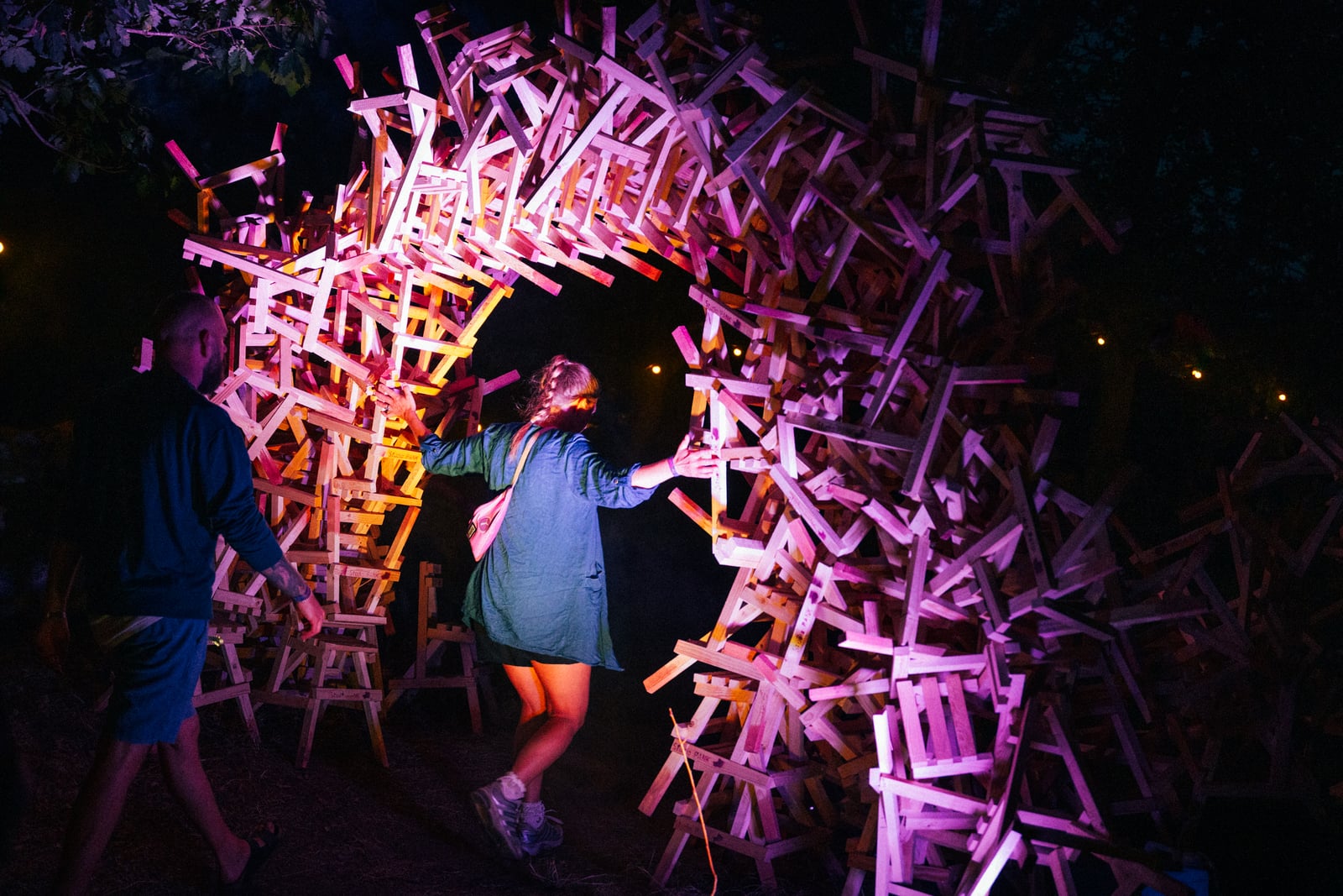 Festival visitors walk through an arch built from stacked wooden chairs, lit magenta against the night forest.