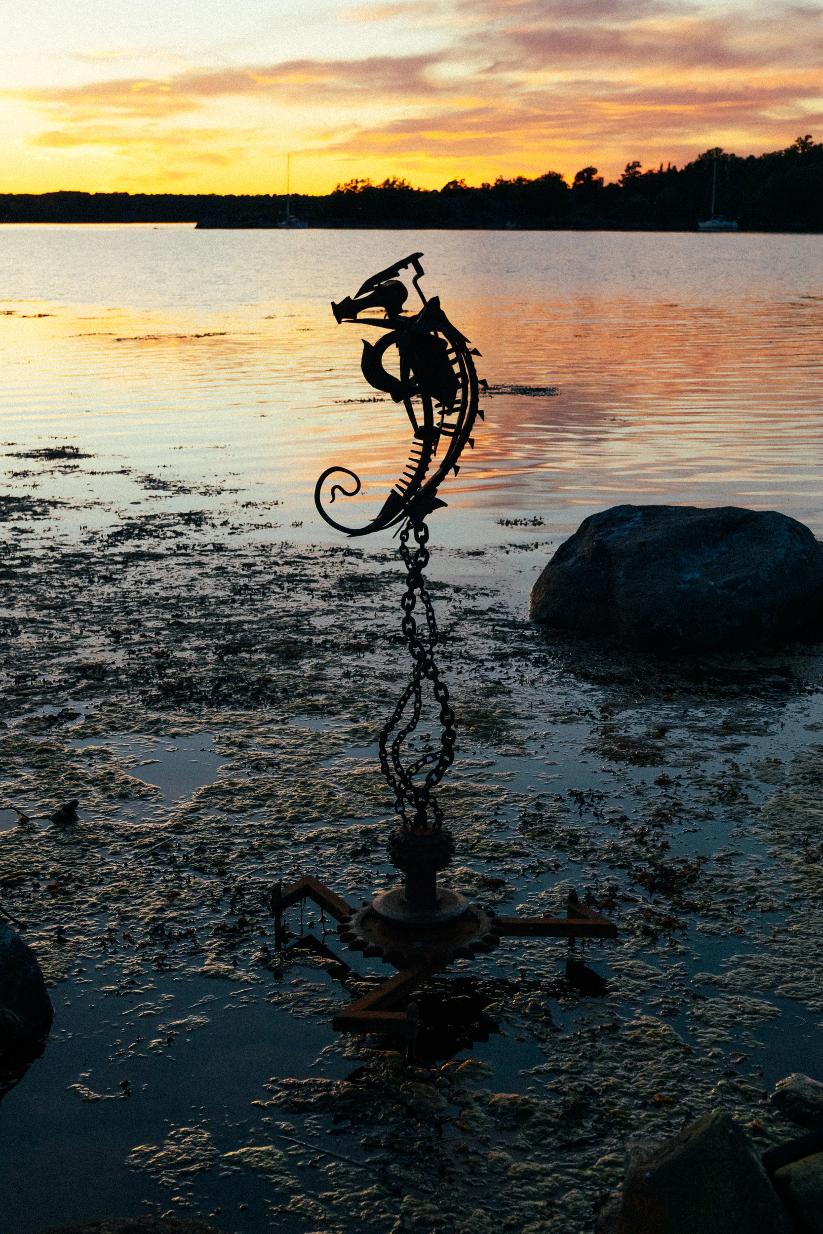 An ironwork seahorse stands in shallow water at sunset, its chain tail running back to the shore.