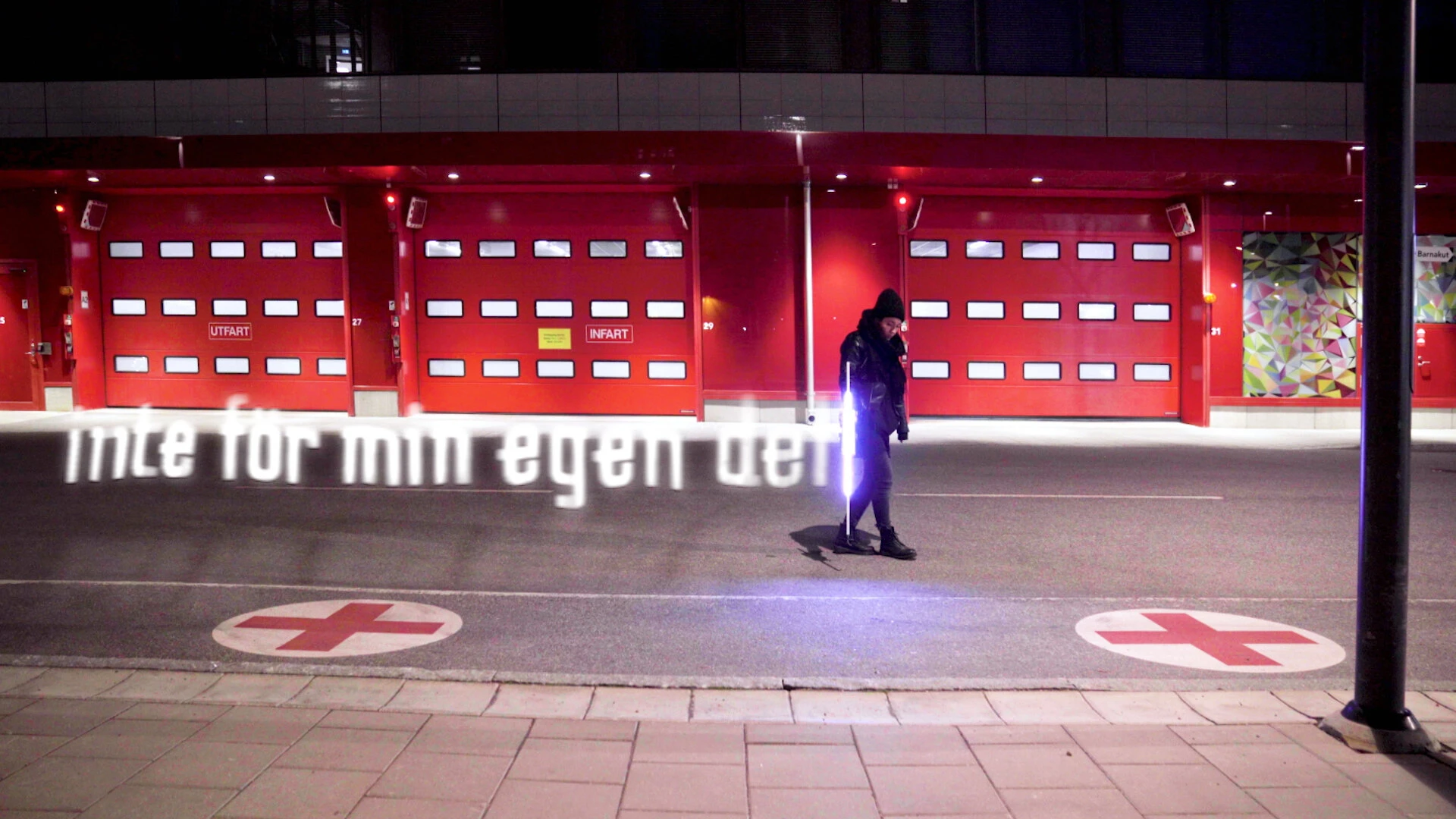 Inte för min egen del, written in light across the forecourt of a red building with garage doors at night.