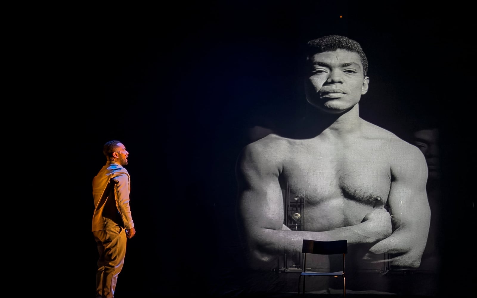 A performer faces a towering black-and-white portrait projected onto the dark stage.