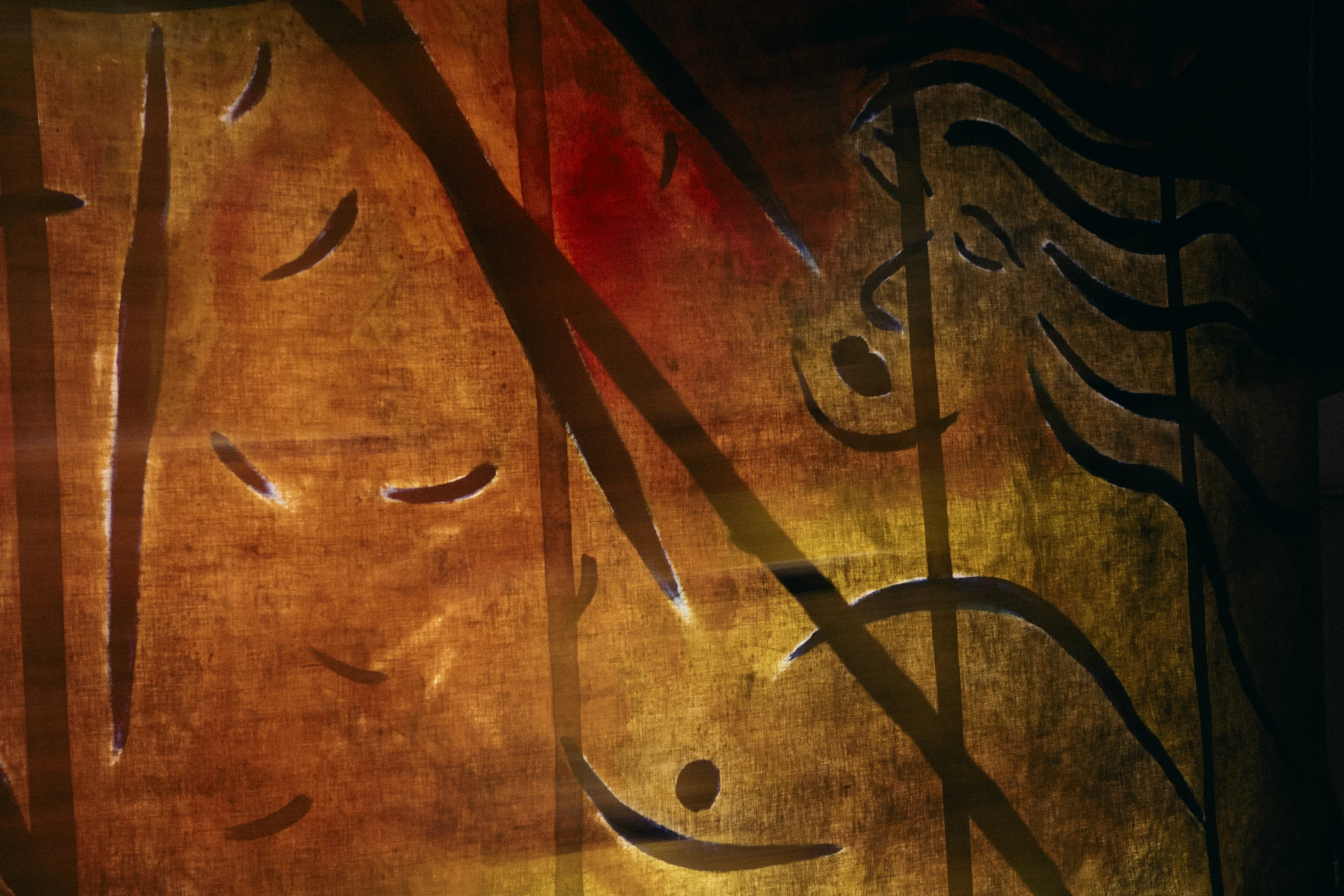 Ochre and red light thrown across a painted surface, its drawn lines and faces catching the projection.