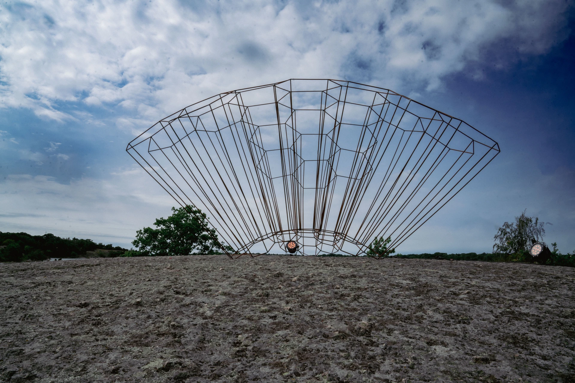 A large fan of thin steel ribs opens against a cloudy sky on the island's bare rock.