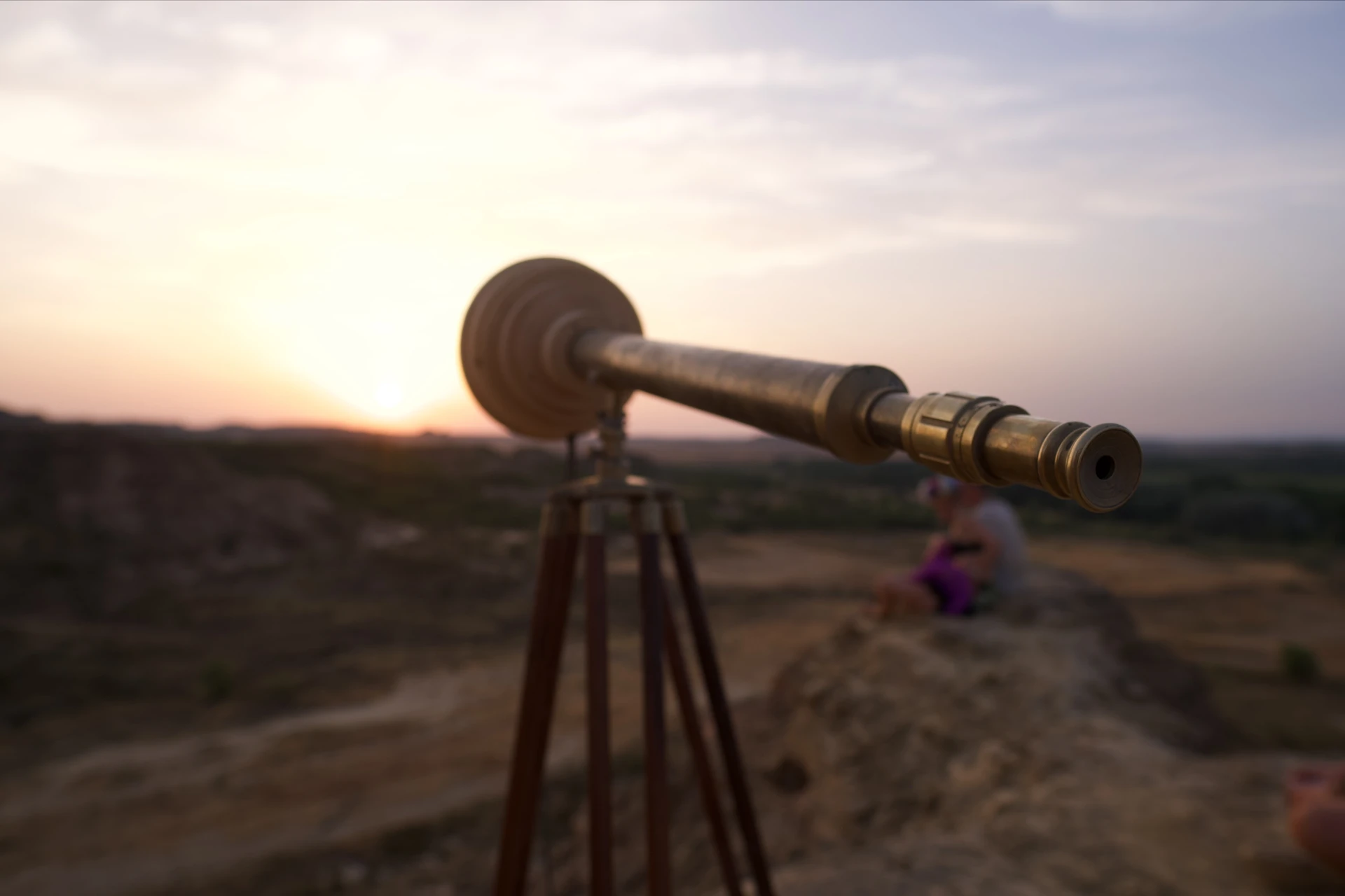 A brass telescope with a circular wooden housing stands on a tripod against a warm desert sunset.