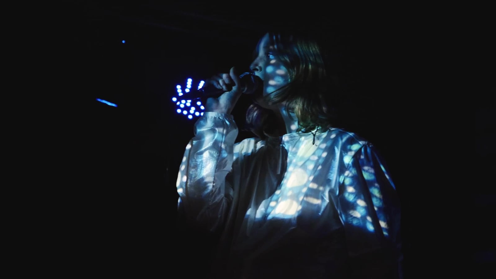 Projected blue and white patterns cover a performer’s face and clothing in a dark audiovisual performance.