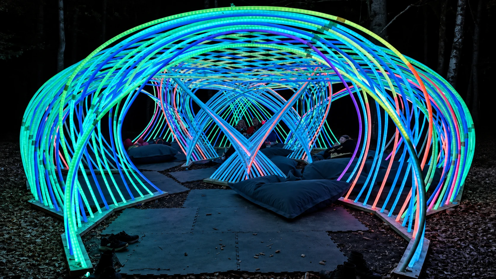 Looking through Kagora's entrance, turquoise and blue arches overlap above people resting on cushions, with pink light deeper inside.