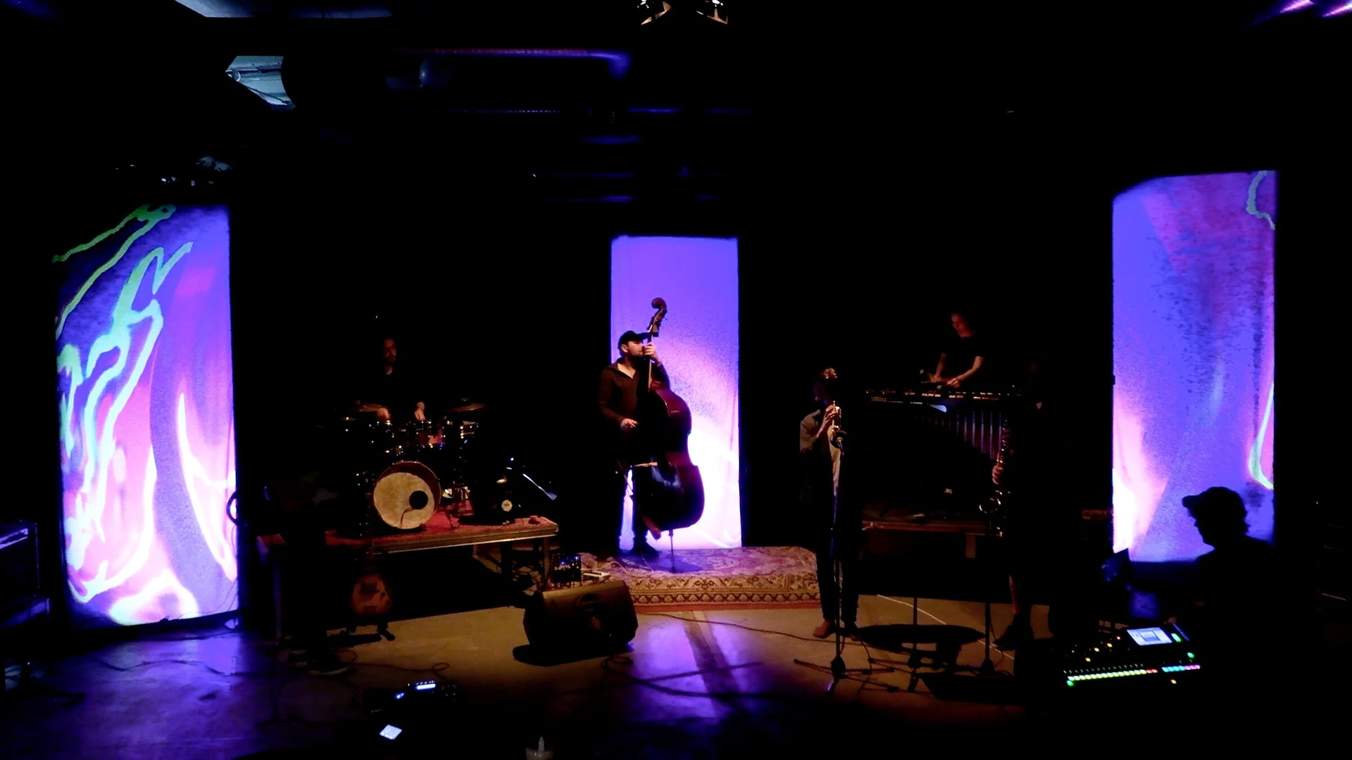 A rehearsal in a dark room with three tall glowing screens of purple and green liquid shapes around the band.