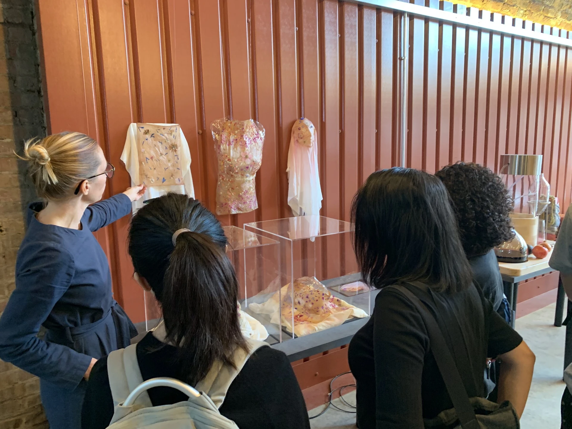 Linda Nurk shows visitors her grown textiles with pressed flowers, hanging above clear vitrines against a rust-red container wall at Open Cell.