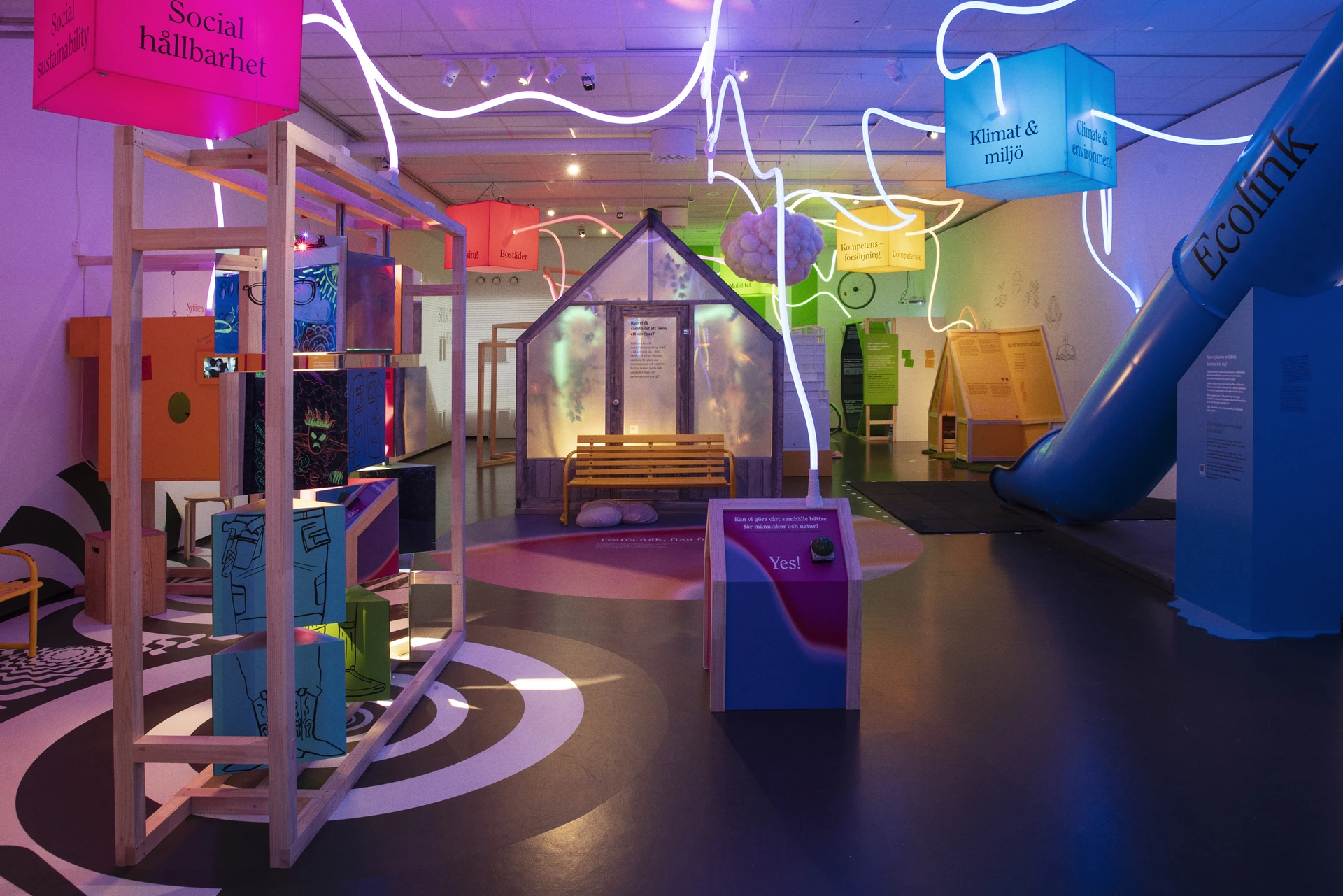 An empty exhibition room with a greenhouse, blue slide and winding overhead lights connecting colourful suspended cubes.