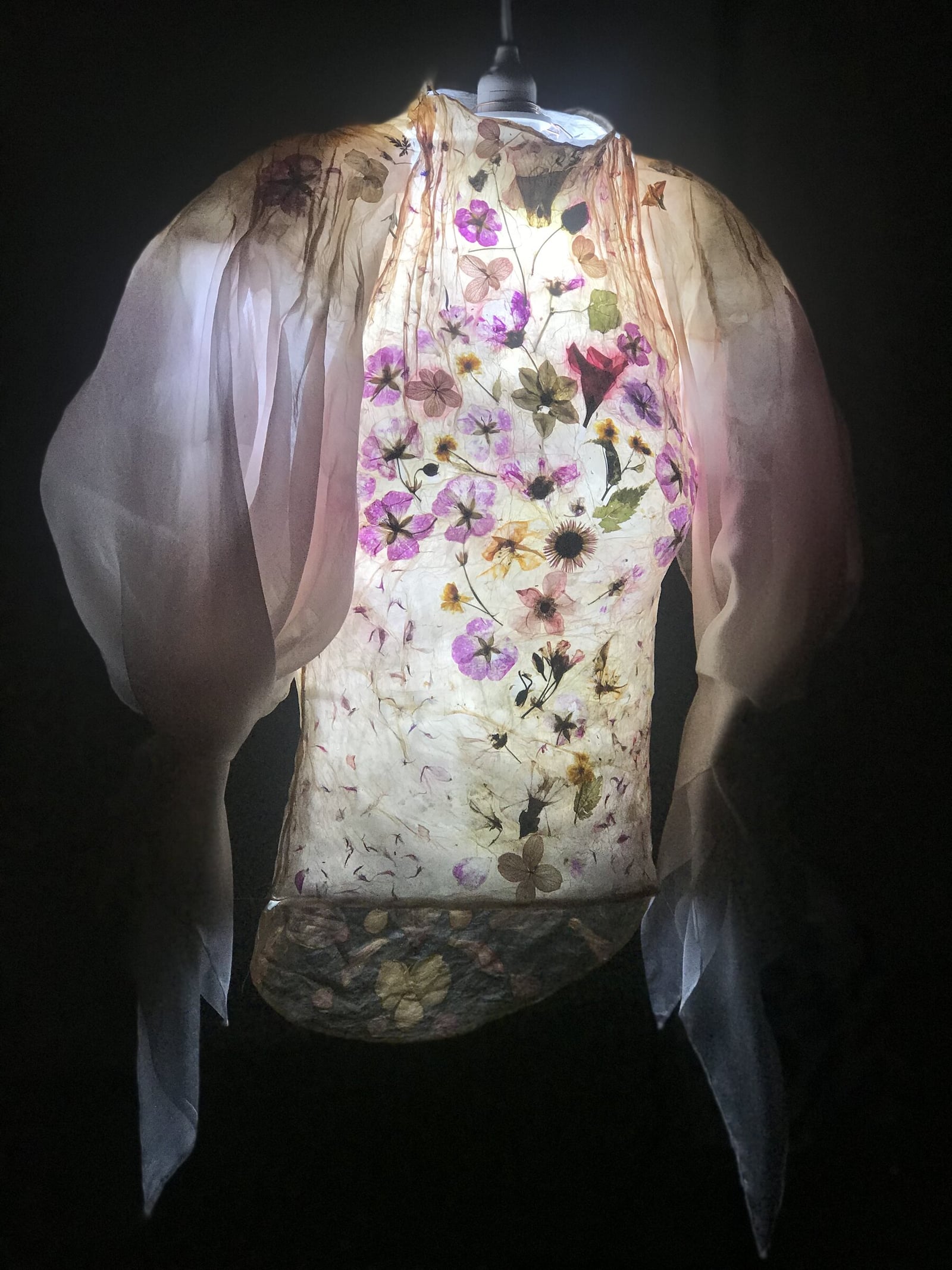 Linda Nurk's Flora bustier lit from inside against black, pressed flowers in pink, purple and yellow set into the grown textile, with sheer sleeves.