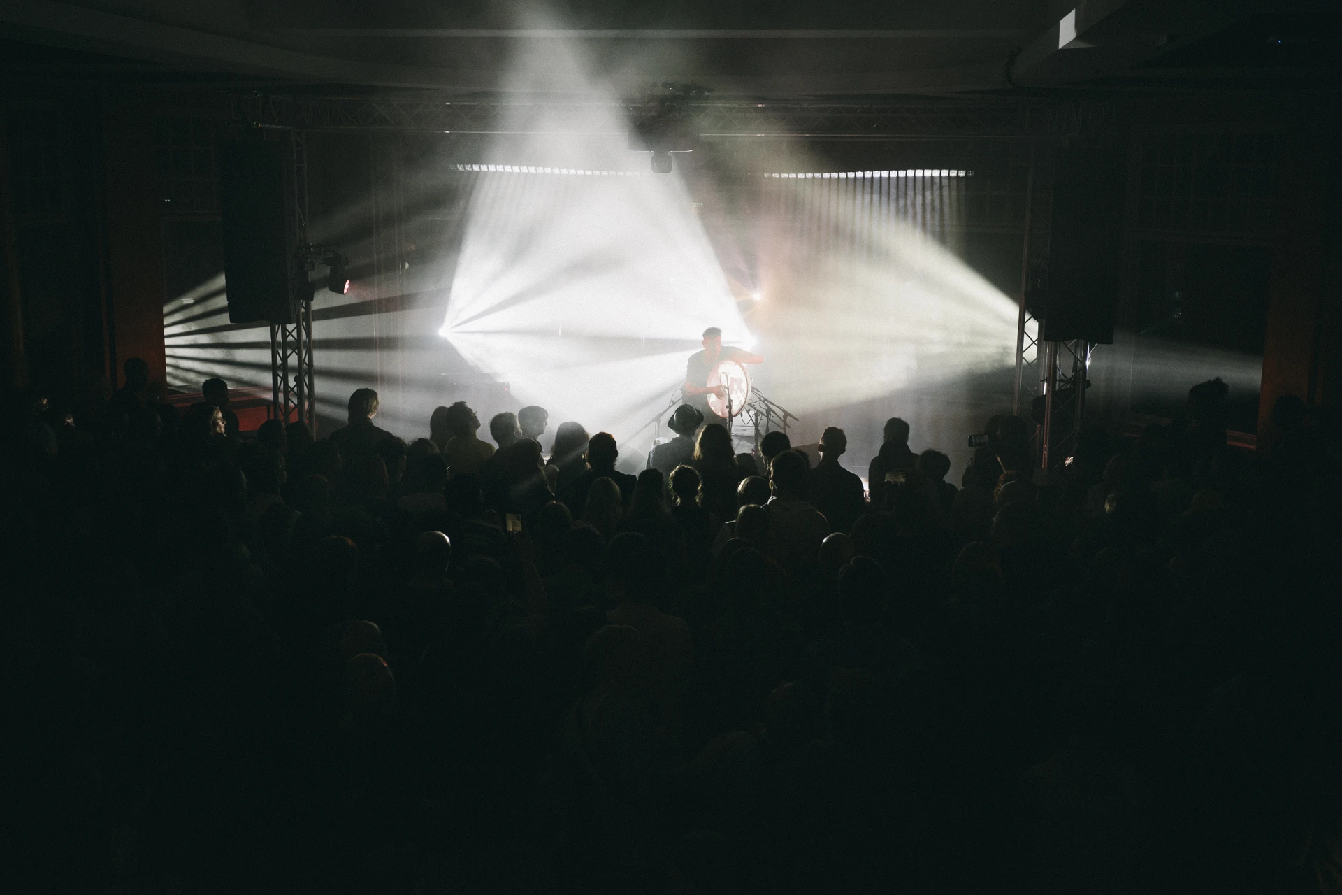 White beams cross above a packed dark hall, the crowd lit only by the stage behind them.