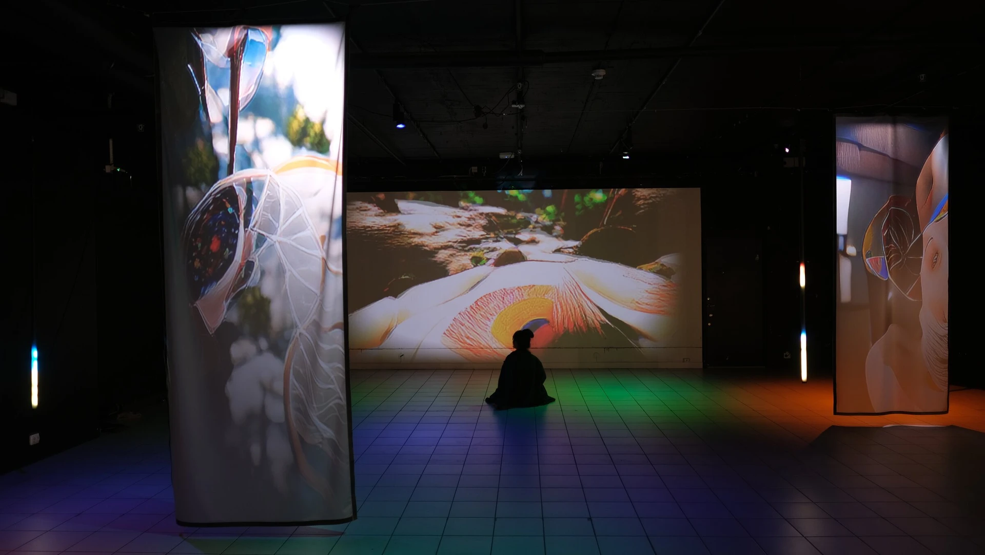 A visitor sits on the floor in front of the wall projection, seen past a scrim, the floor washed in blue and green.