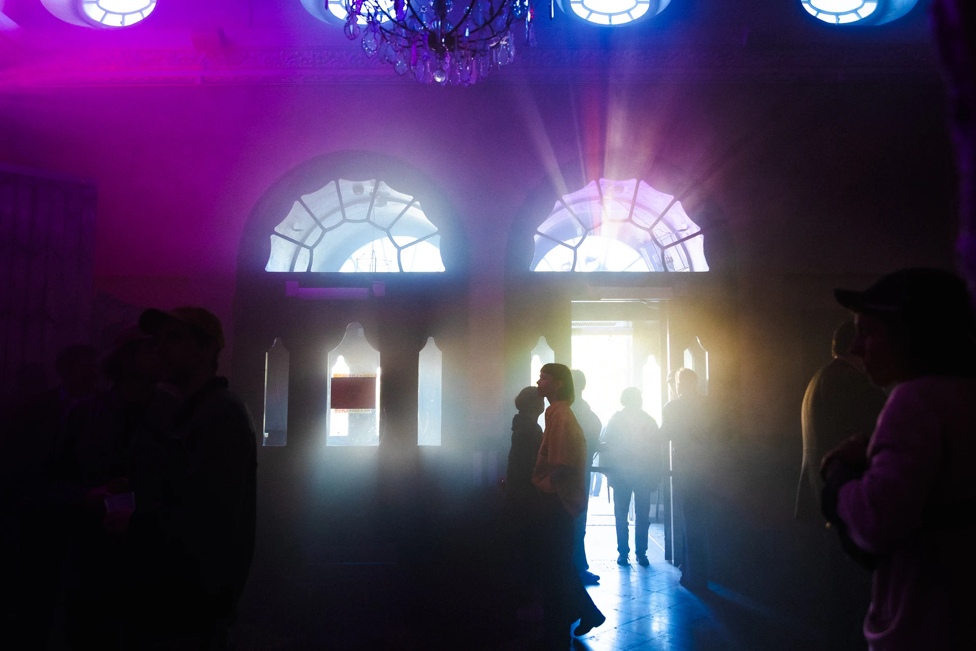 Arched museum windows glow magenta and blue above a crowd in silhouette, a bright doorway at the centre.