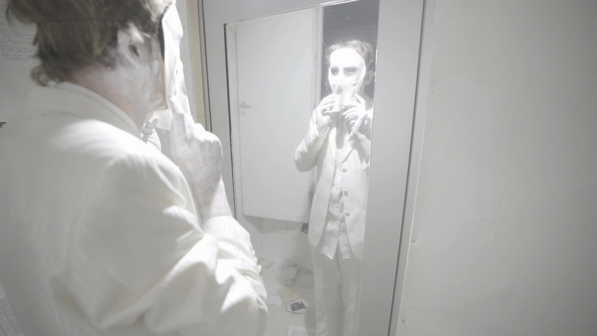 The man in the white suit faces a doorway mirror; the reflection looking back wears a white mask.