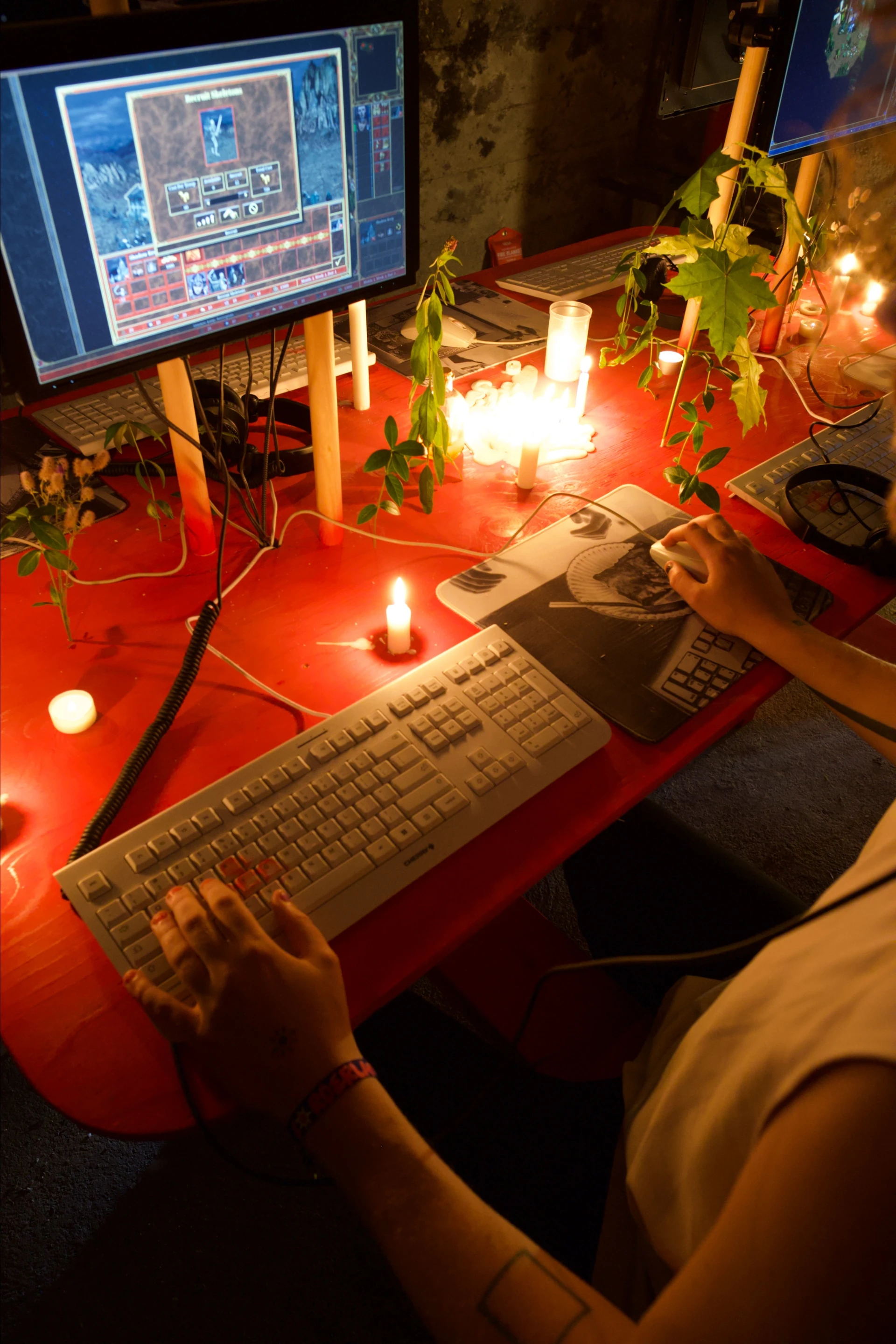 An overhead view of a player's hands at the keyboard and mouse, with Heroes of Might and Magic III on the screen and candles between the stations.