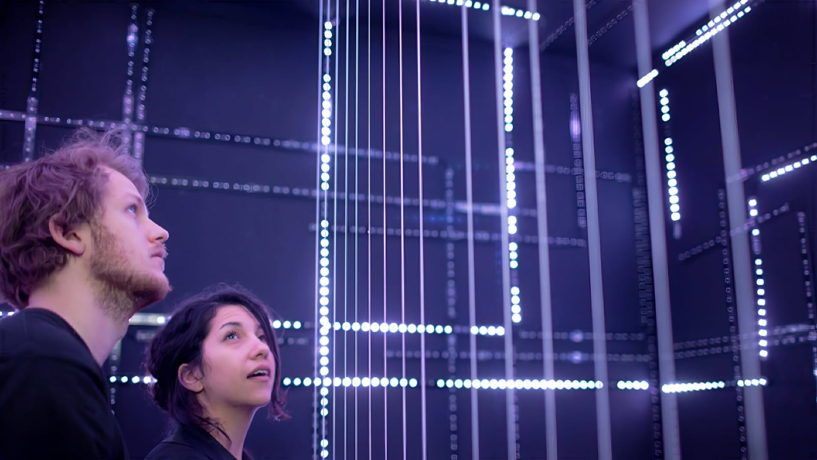 Two visitors look up through a grid of glowing vertical strings in a dark room.
