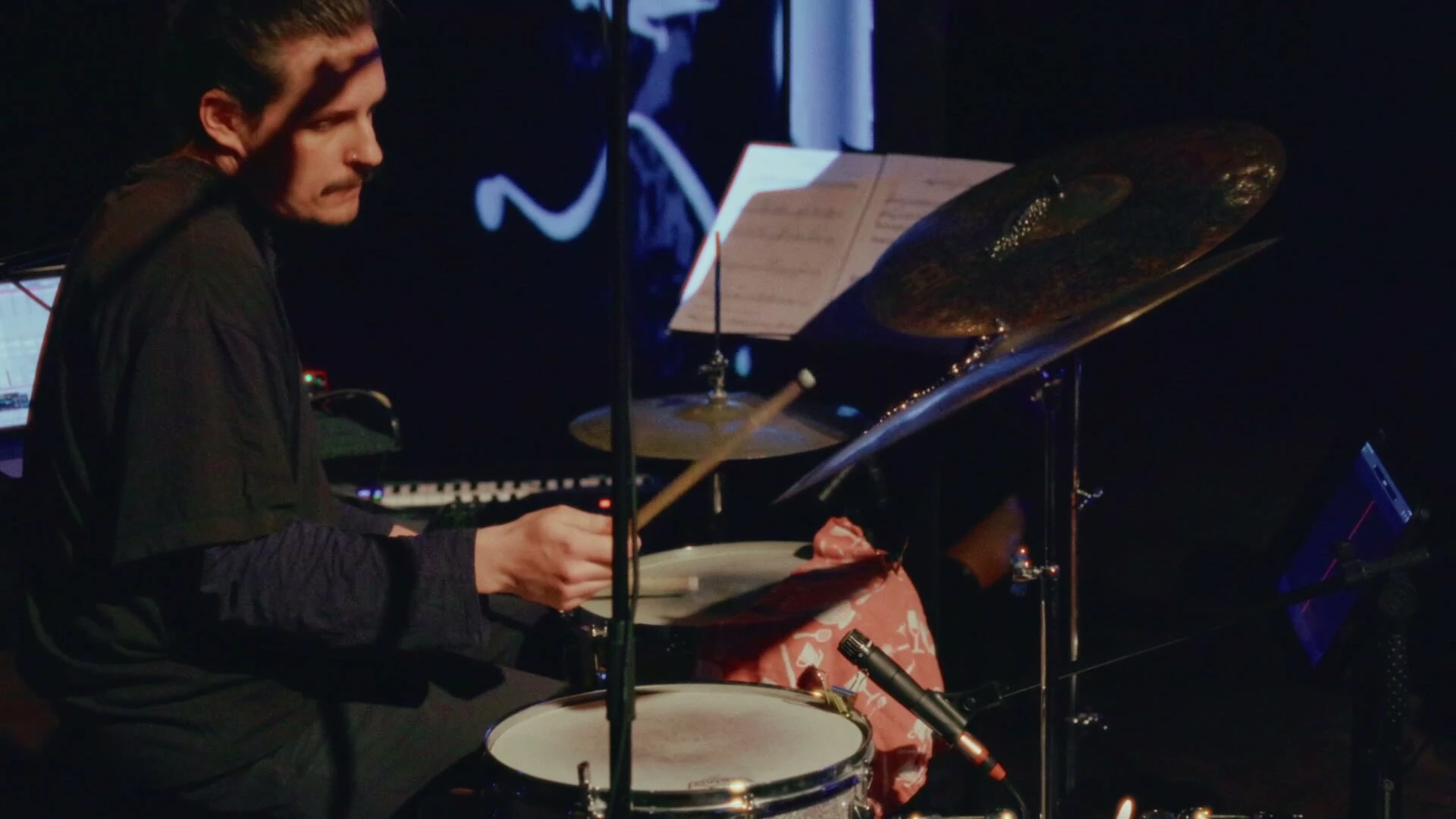 The drummer at his kit, sheet music on the stand, with a white stroke of the projected score above.