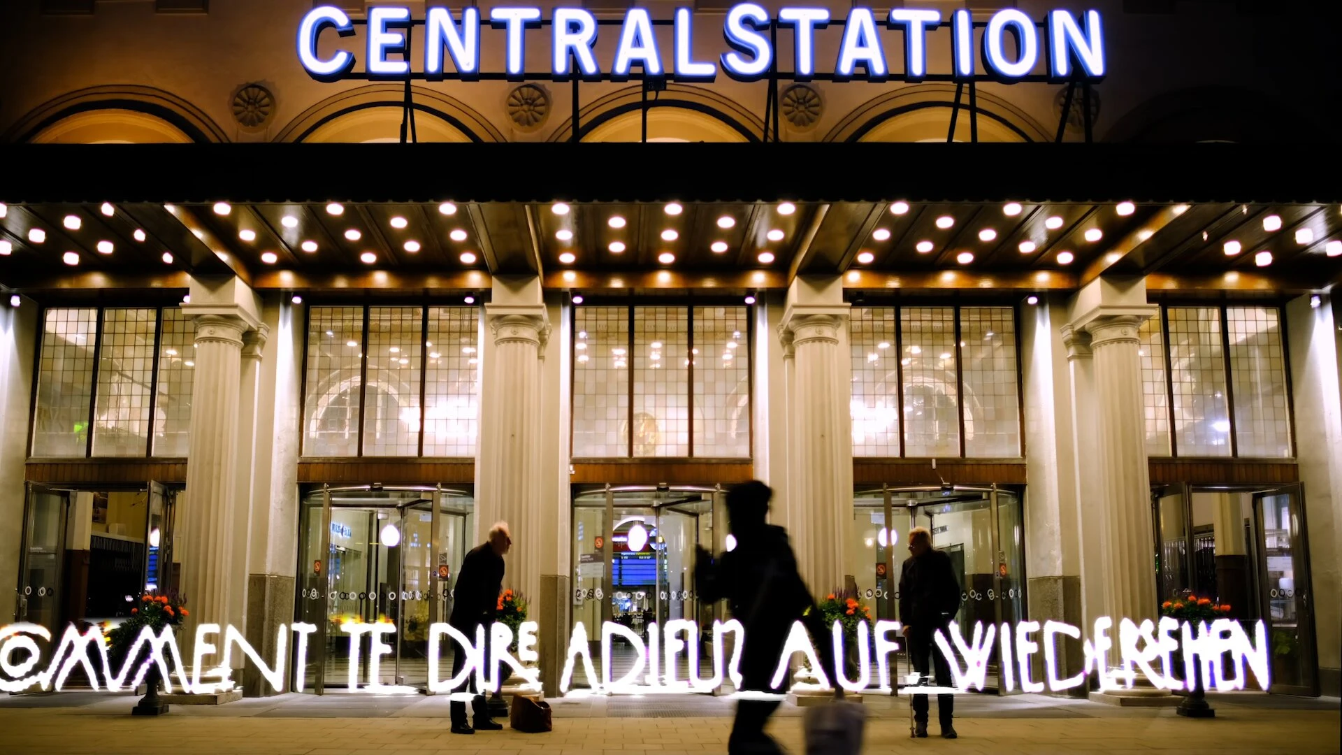 Comment te dire adieu, auf Wiedersehen, written in light along the entrance of Stockholm Centralstation as travellers pass.