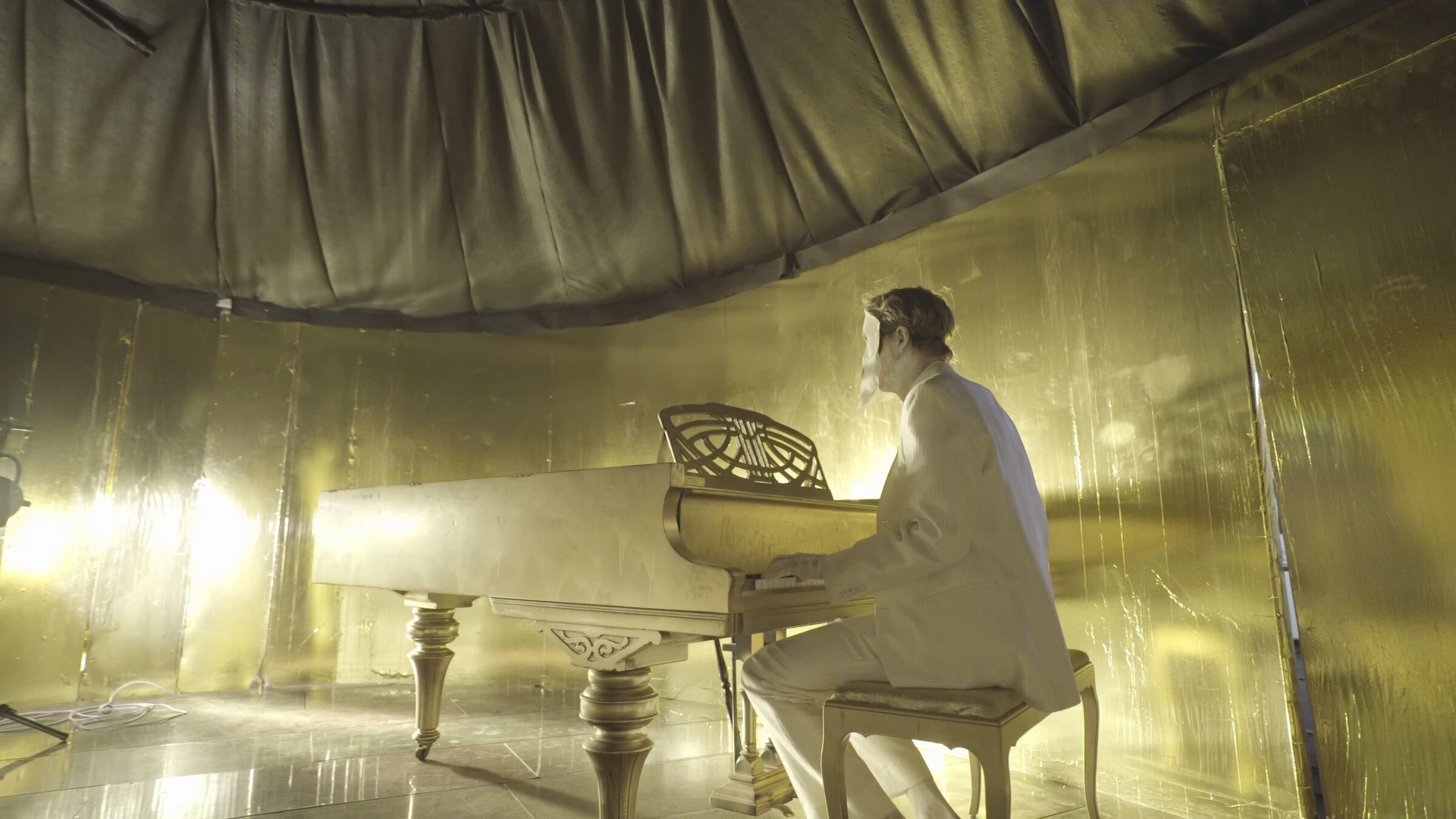 The masked figure plays a grand piano in a room lined with gold foil, lit by lamps on the floor.