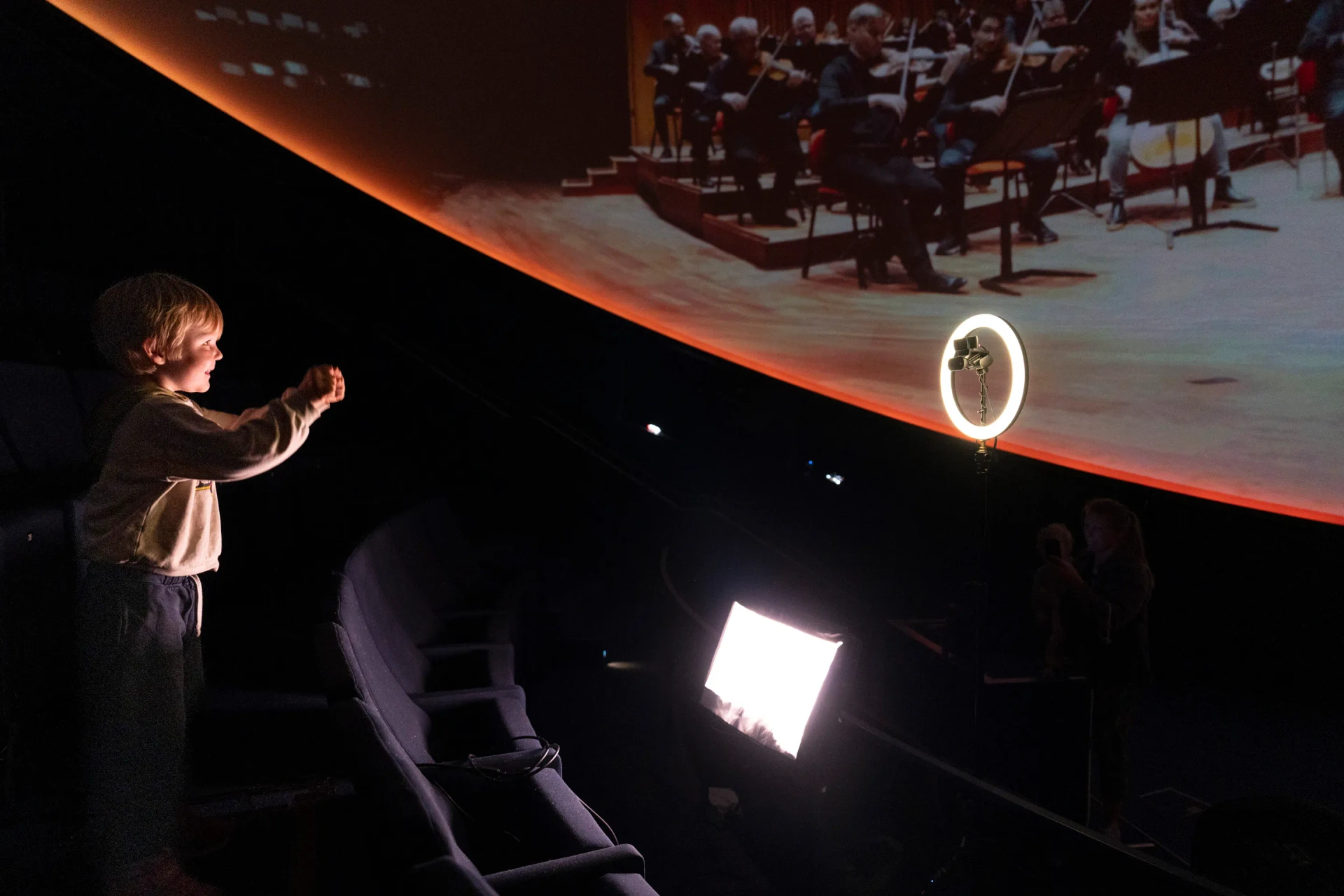 A child raises both arms beneath the projected orchestra, facing a tracking camera mounted inside a ring light.