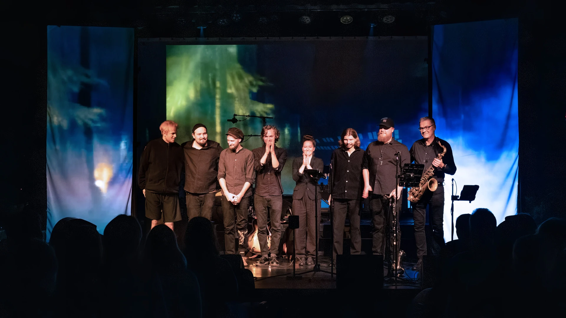 The musicians take a bow under misty projections of a pine forest in green and blue.