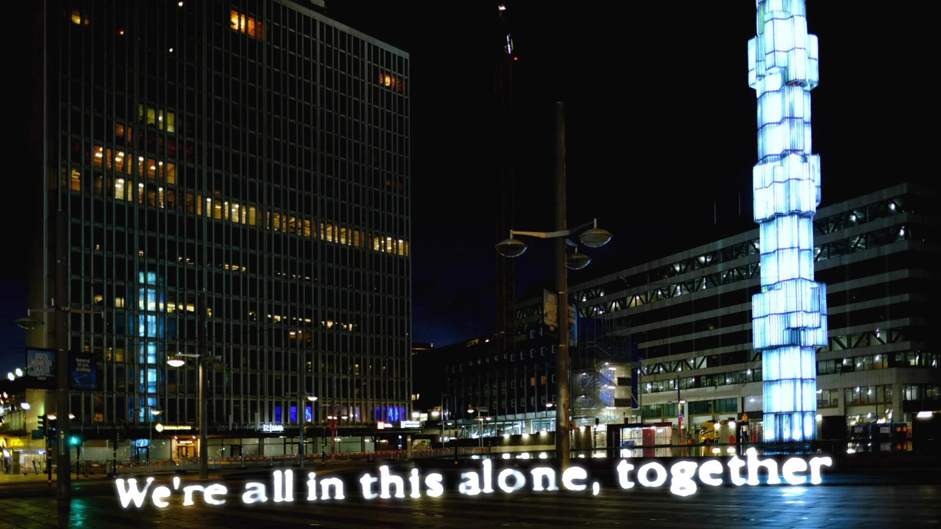 We're all in this alone, together, written in light at Sergels torg beside the lit glass obelisk.