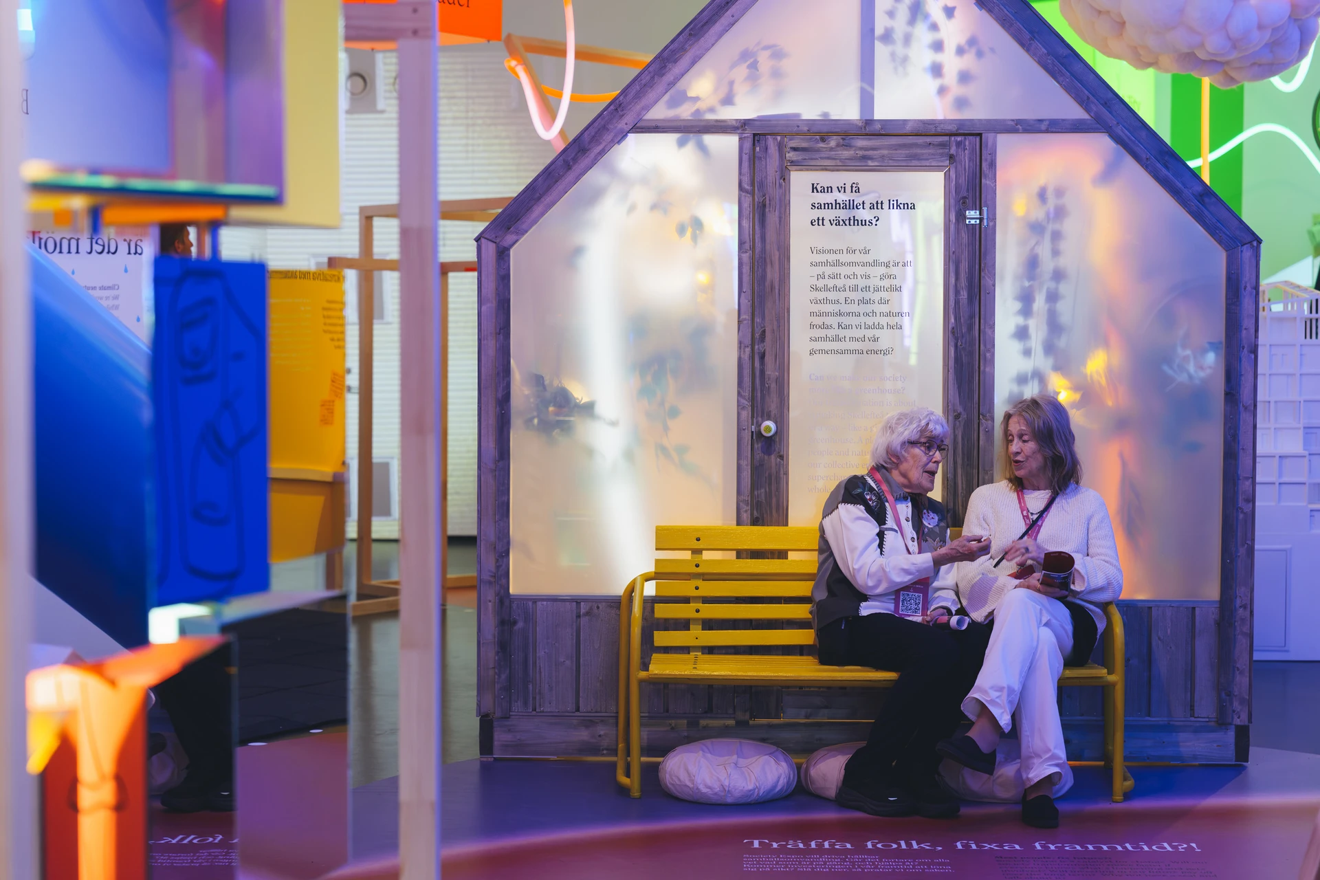 Two visitors sit on a yellow bench inside the translucent greenhouse beneath the suspended cloud and winding lights.