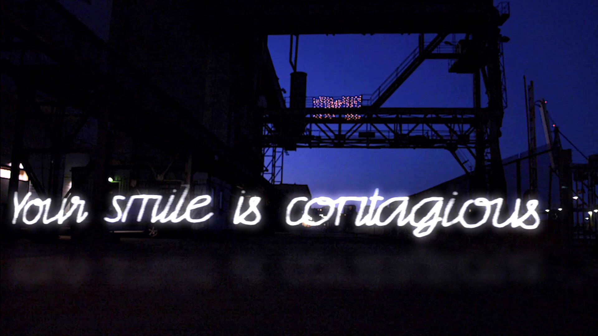 Your smile is contagious, written in white script light beneath a dark steel structure against a blue night sky.
