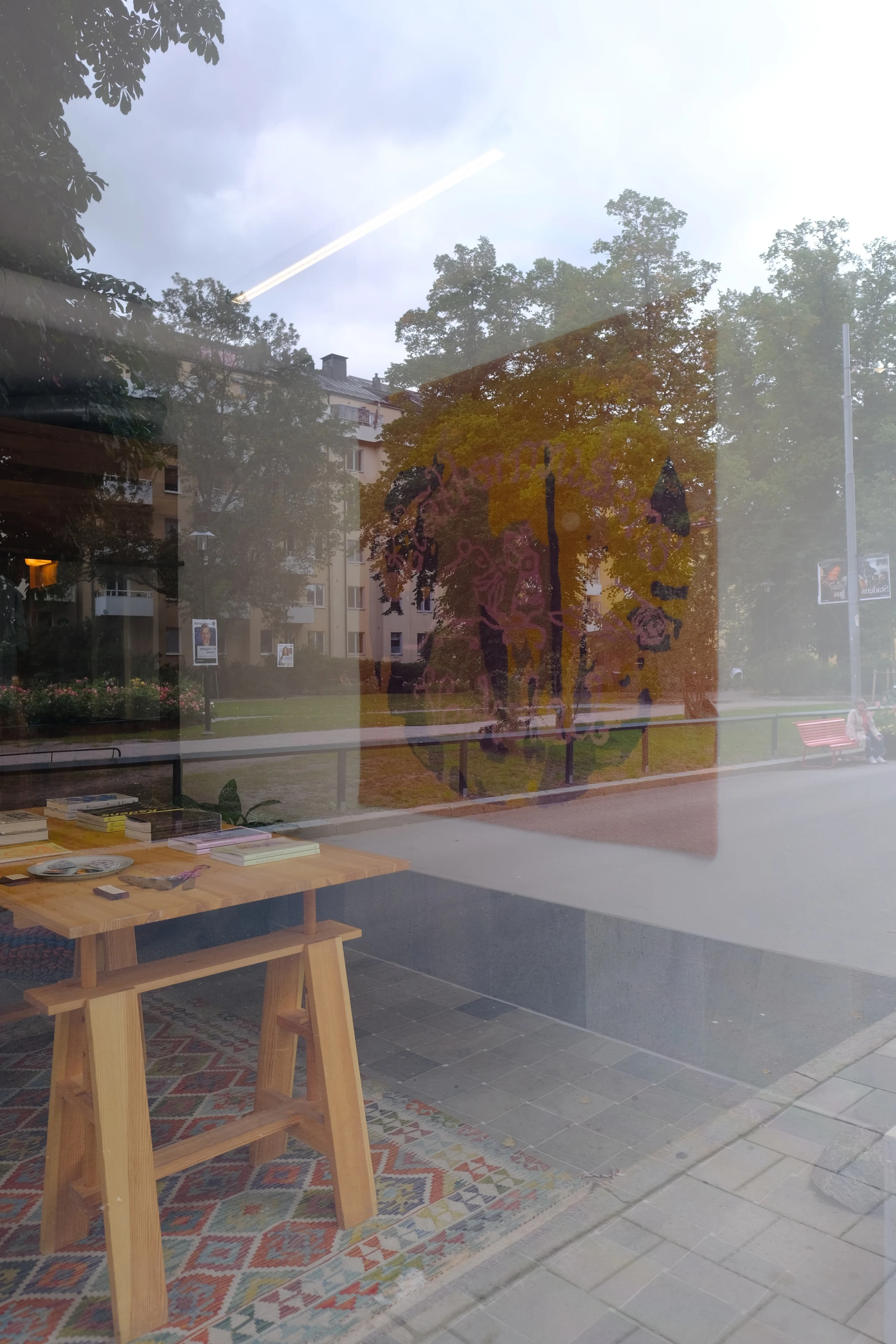 The rug and workshop table are visible through the shopfront glass, layered with reflections of trees and the street.