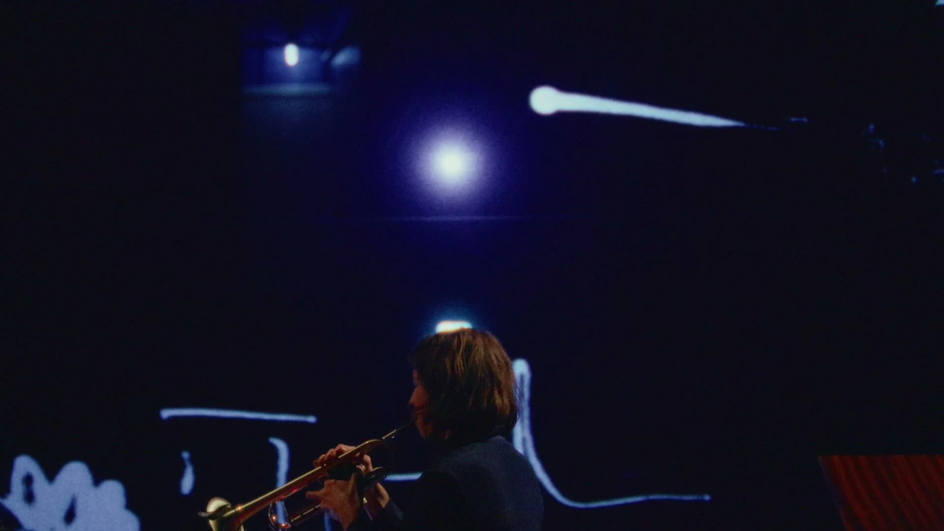 Adam Sass plays trumpet in blue light, a white line of the score curling across the wall behind him.