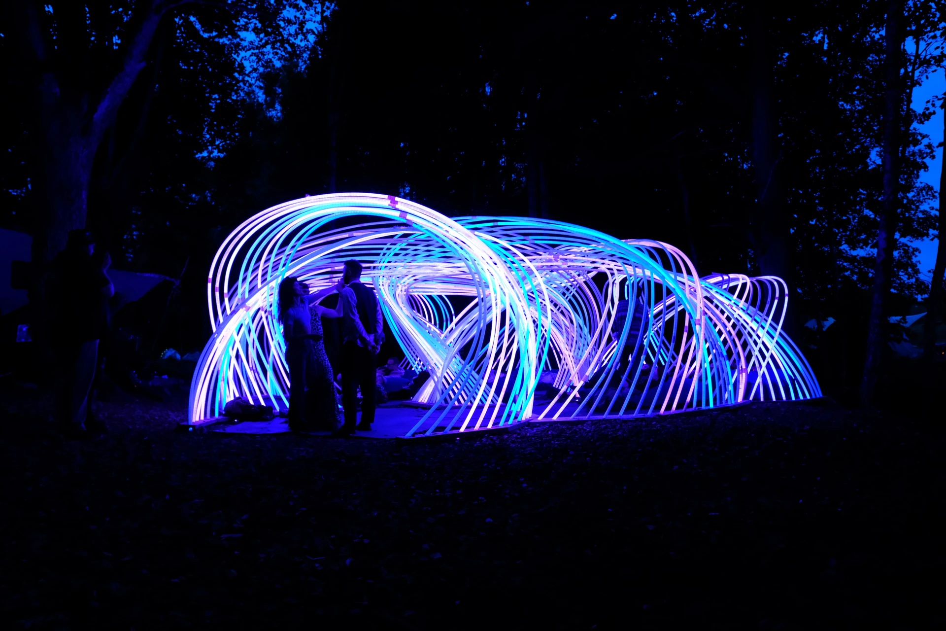 Visitors stand beneath Kagora's overlapping blue, violet and white light arches among trees at dusk.