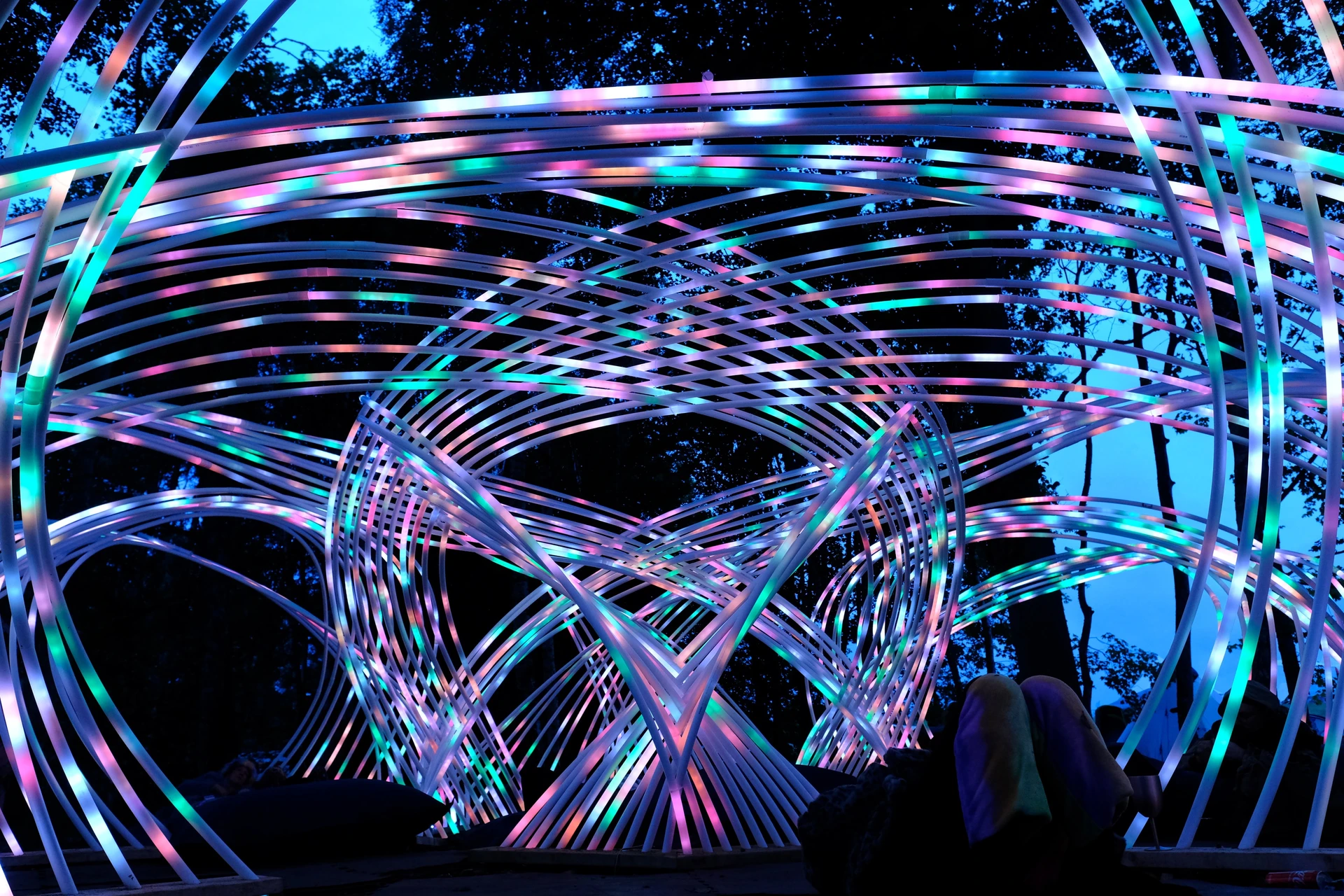 Looking up from inside Kagora, multicoloured light curves cross above visitors beneath the evening sky.
