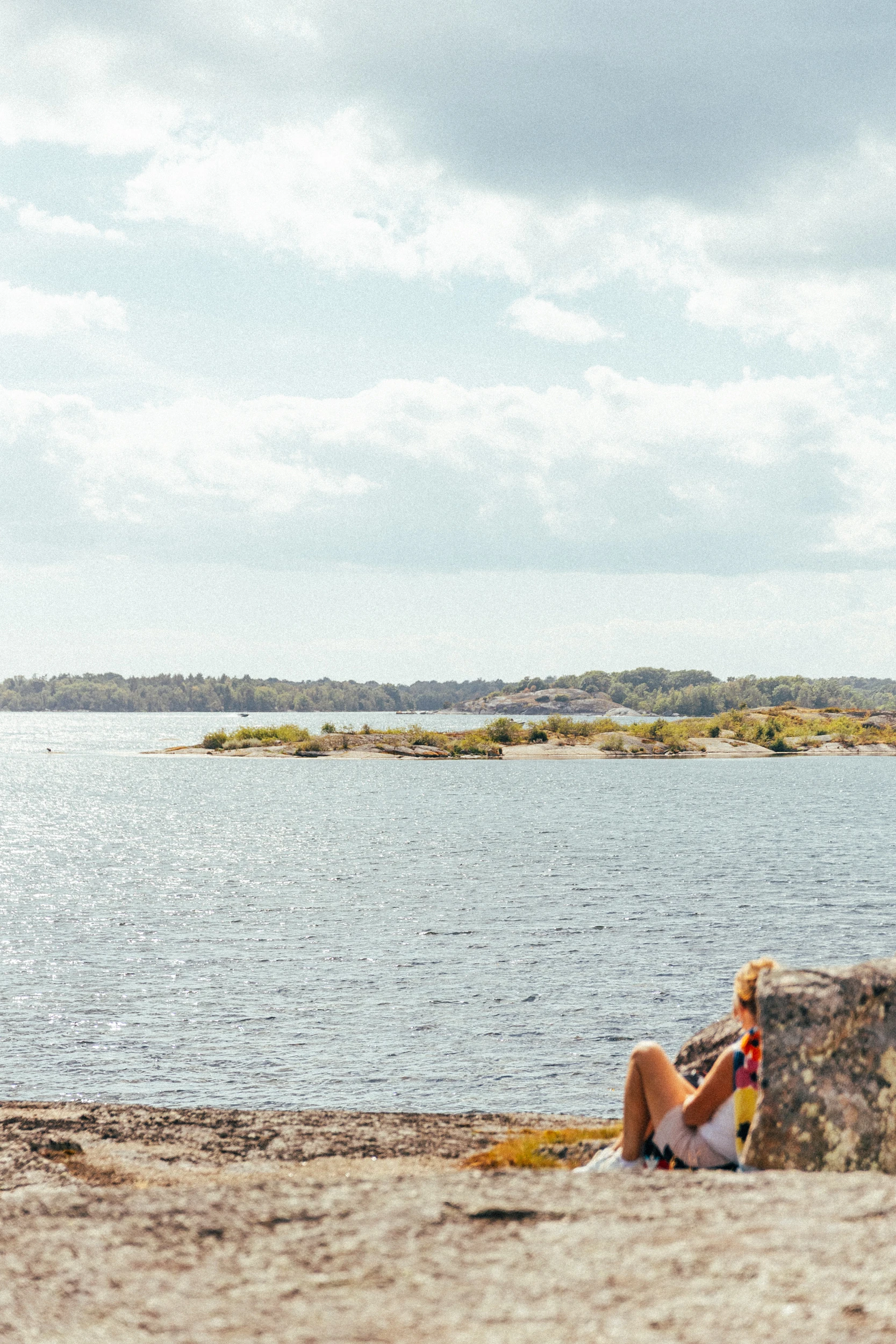 Someone sits alone on the shoreline rock looking out at the low islands of the archipelago.