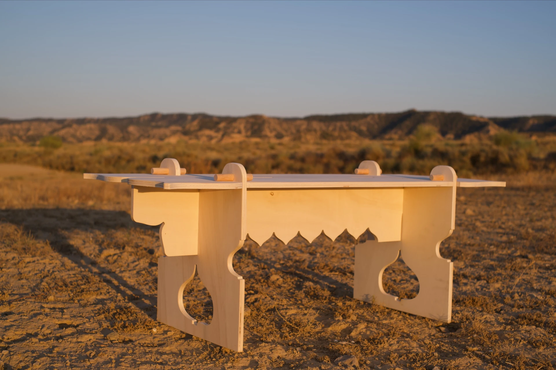 Rose Hallgren's wooden bench stands in the desert in warm evening light, with curved cutouts in its legs and a scalloped edge beneath the seat.