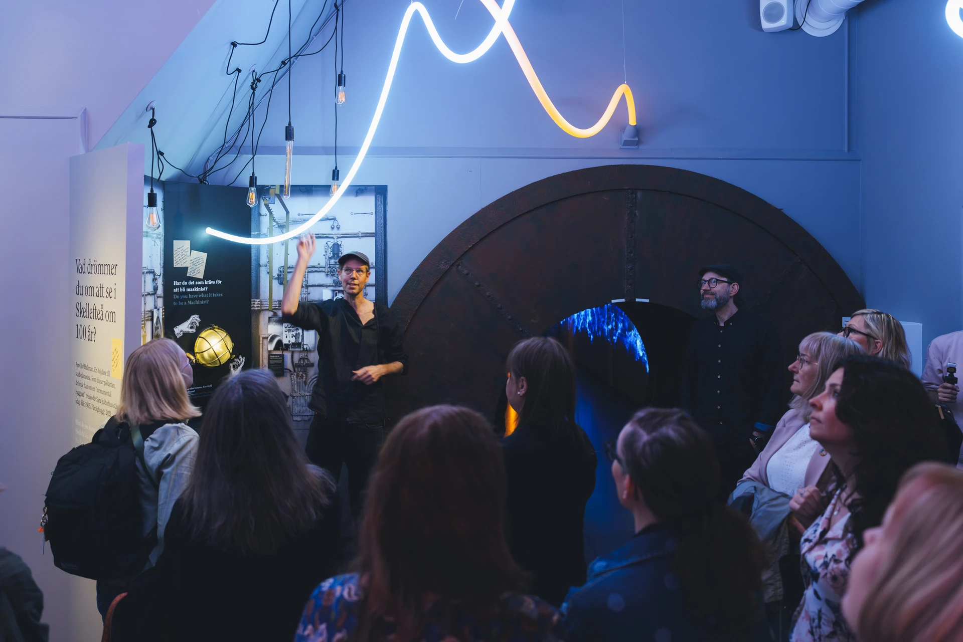 A group gathers beneath a curving blue and amber light line beside the round wooden hydropower tunnel.