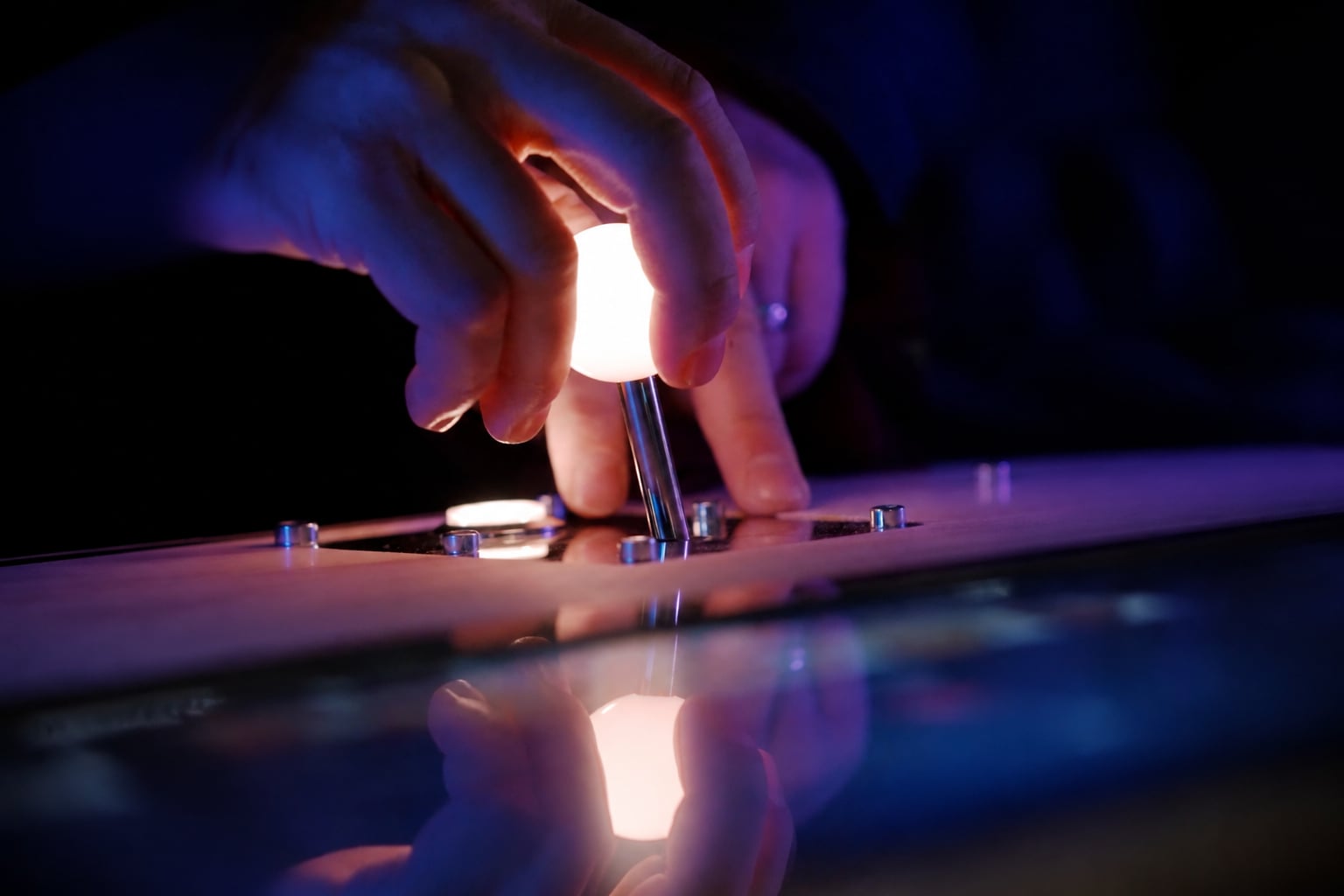 A hand grips a glowing arcade joystick under blue and purple light, reflected in the controller’s glossy surface.