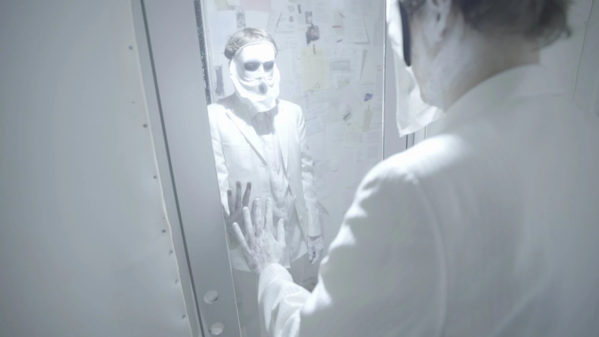 With the light on, the man and his masked reflection press their hands against the glass.