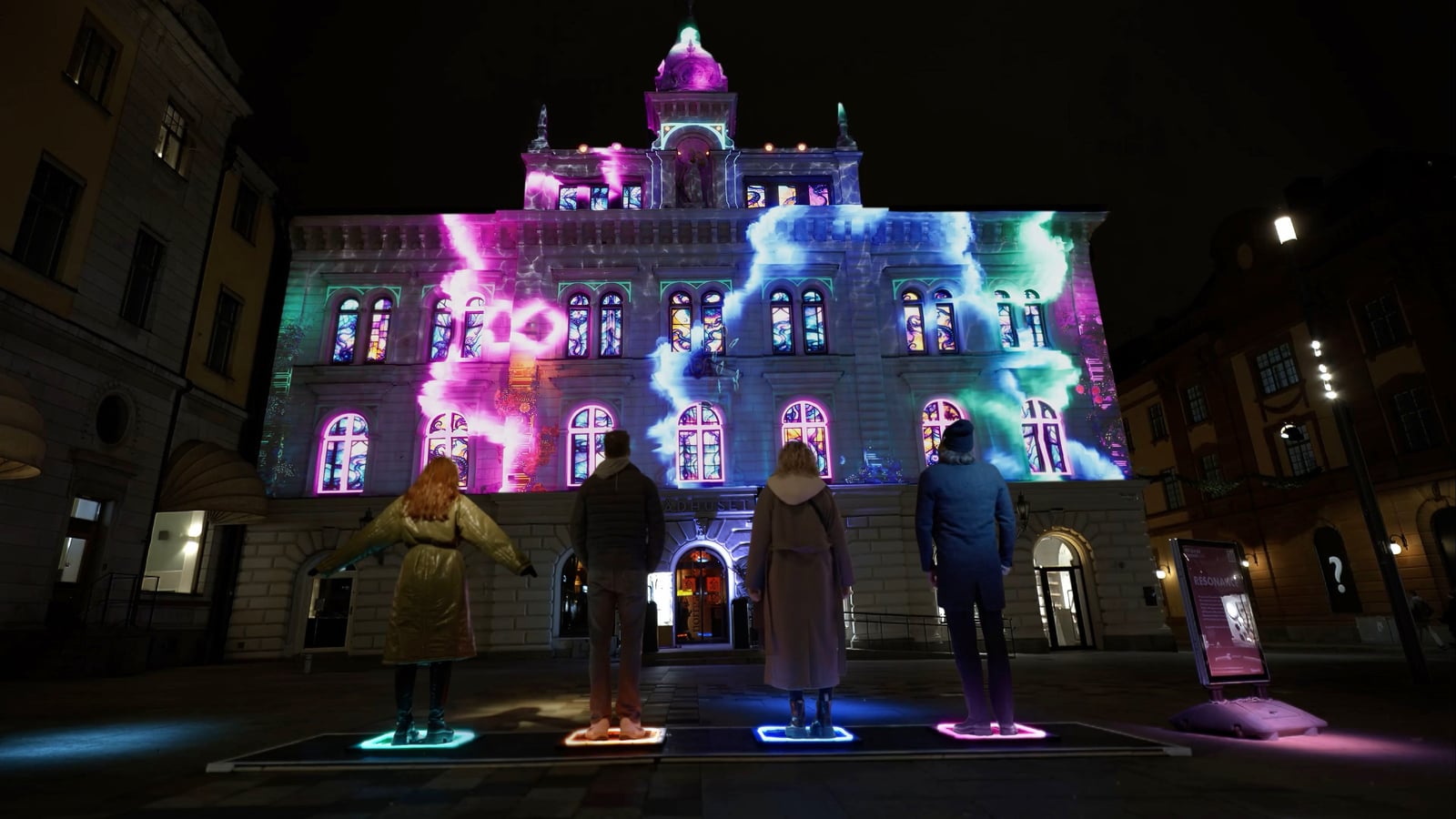 Purple, blue and green projections cover Uppsala Town Hall while people gather around a controller in the square.