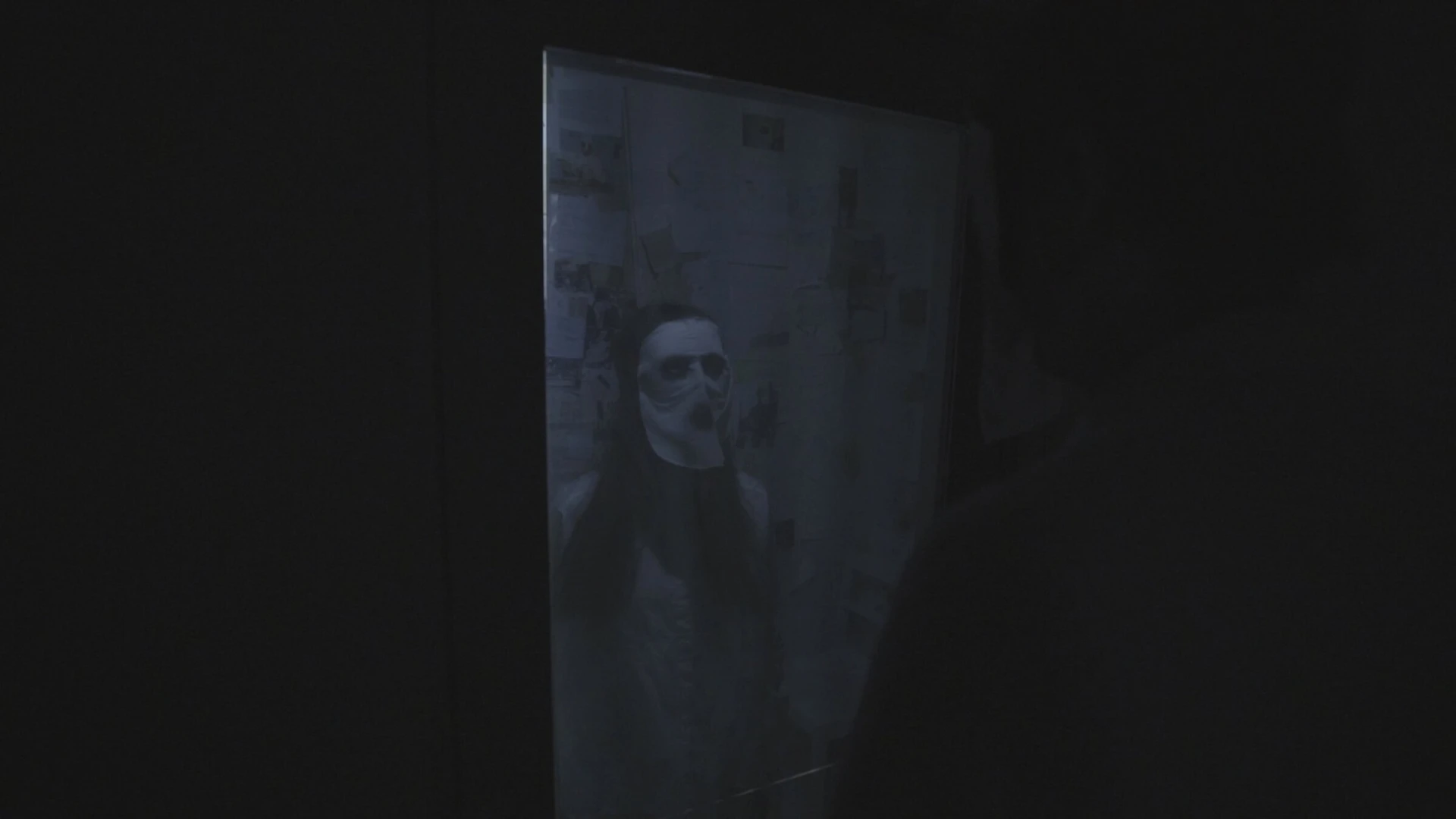 In darkness, a masked face shows faintly through the glass of the door.