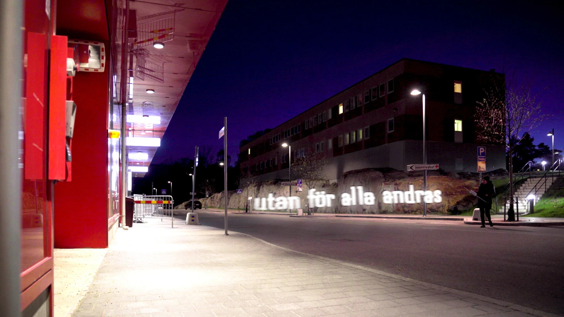 Utan för alla andras, written in light along a pavement beside the same red building.