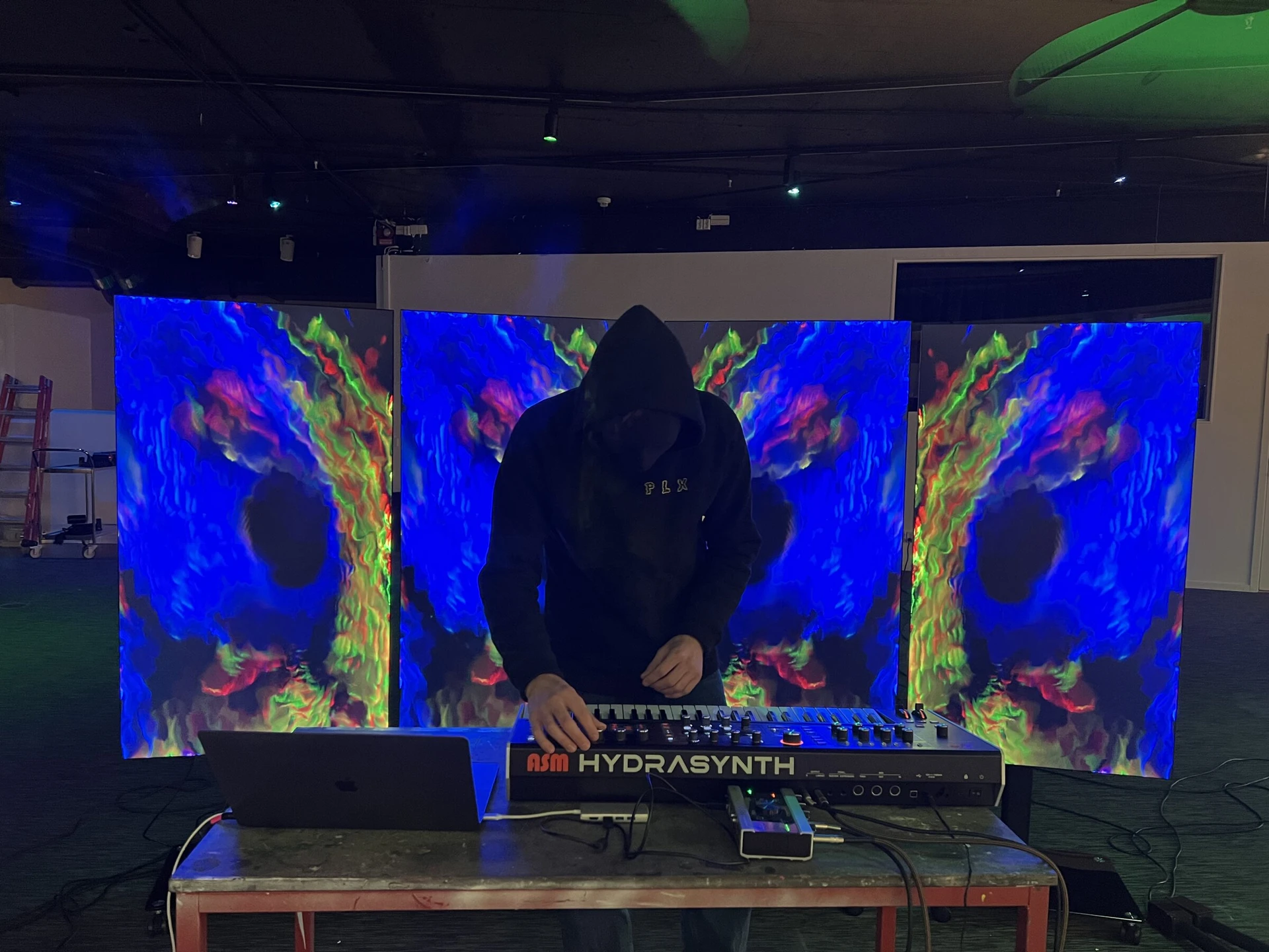 A hooded, masked performer plays a Hydrasynth synthesiser in front of three screens of flowing blue, green and red patterns.