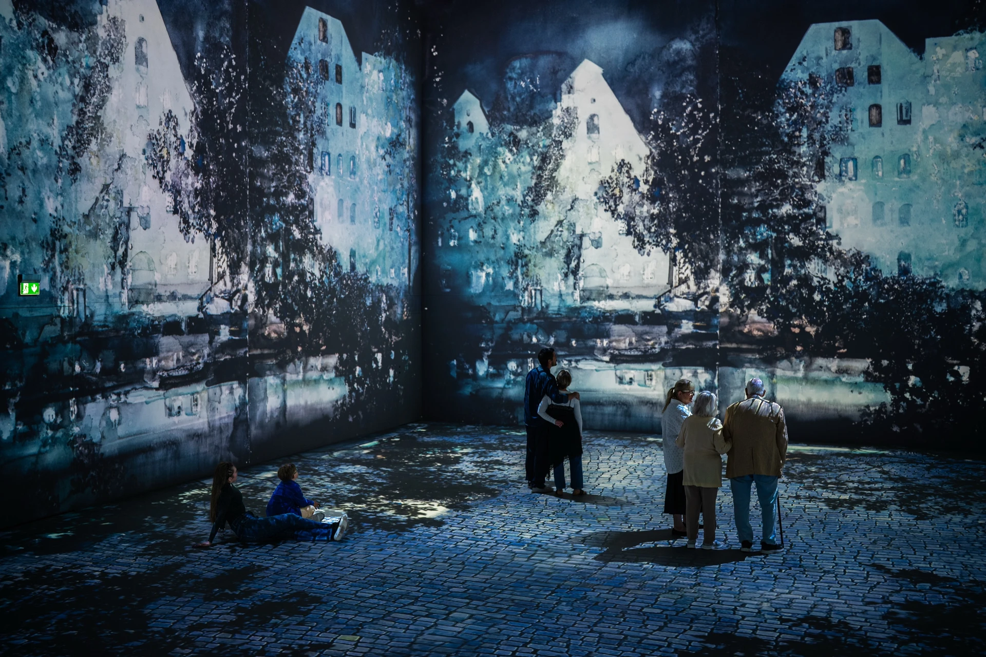 Two visitors sit on the cobbled floor projection while others stand in the corner of the hall among blue watercolour houses.