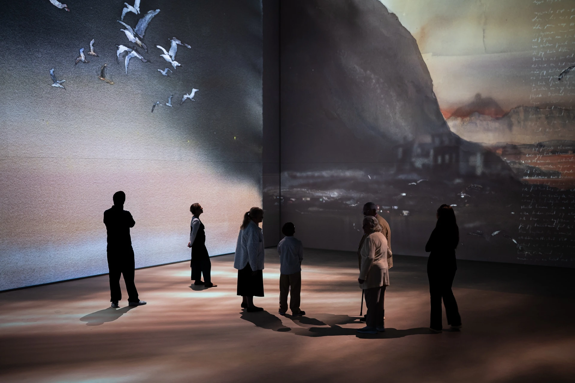 Visitors stand as silhouettes on a warmly lit floor while gulls fly across a projected Lofoten sky and mountain.