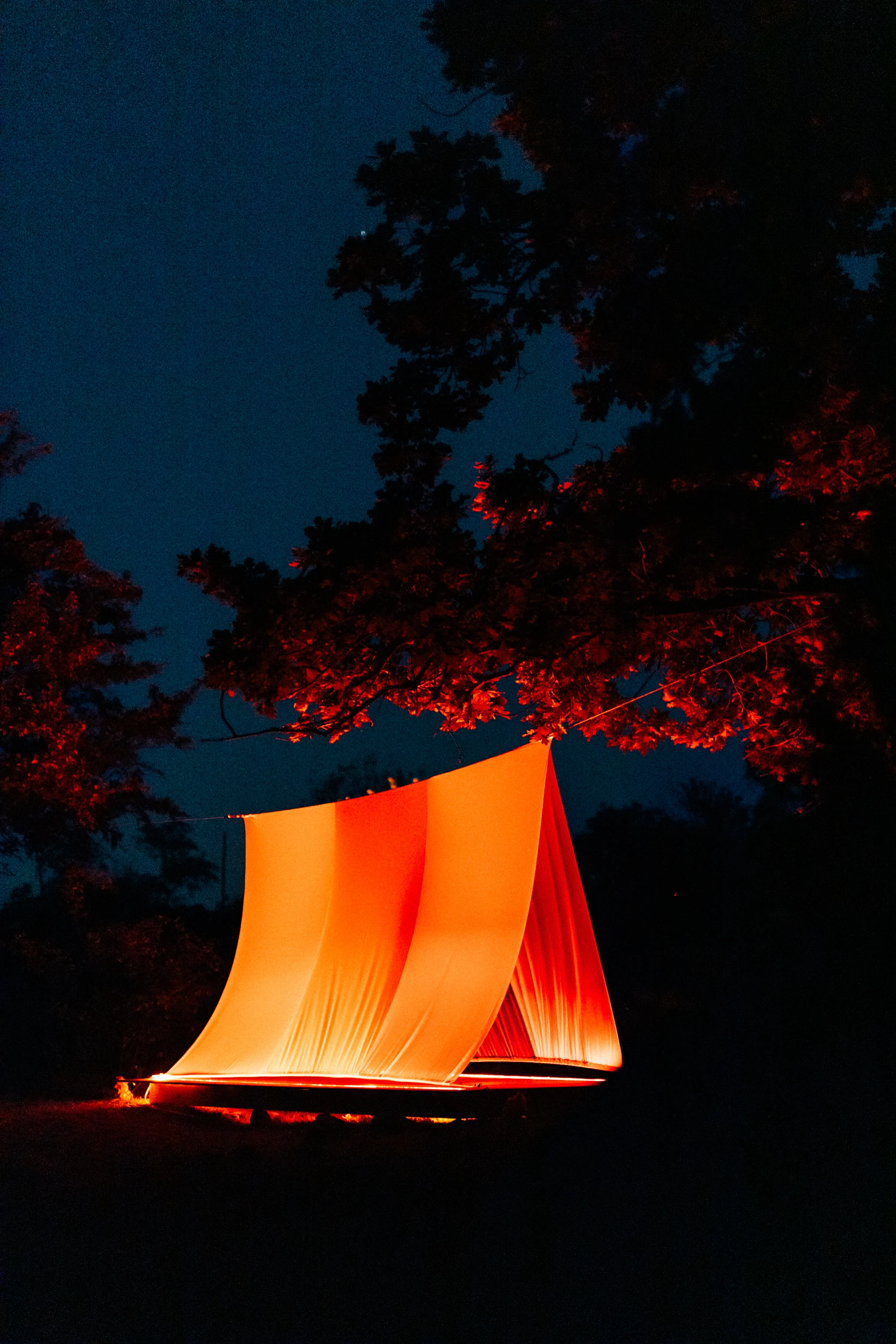 A tall sail of fabric stretched between poles in the dark, lit orange from below, with red light in the trees behind it.