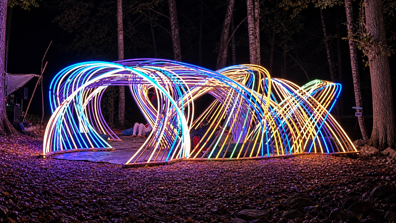 Kagora's illuminated arches spread across a woodland clearing, with white, cyan and yellow light tracing the woven pavilion.
