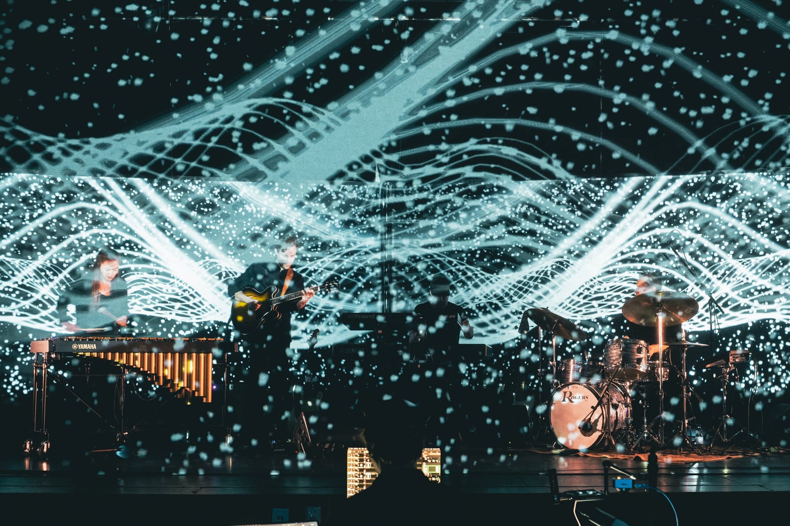 Waves of white particles fill a gauze and the screen behind it, with a vibraphonist, guitarist and drummer playing between the two layers.