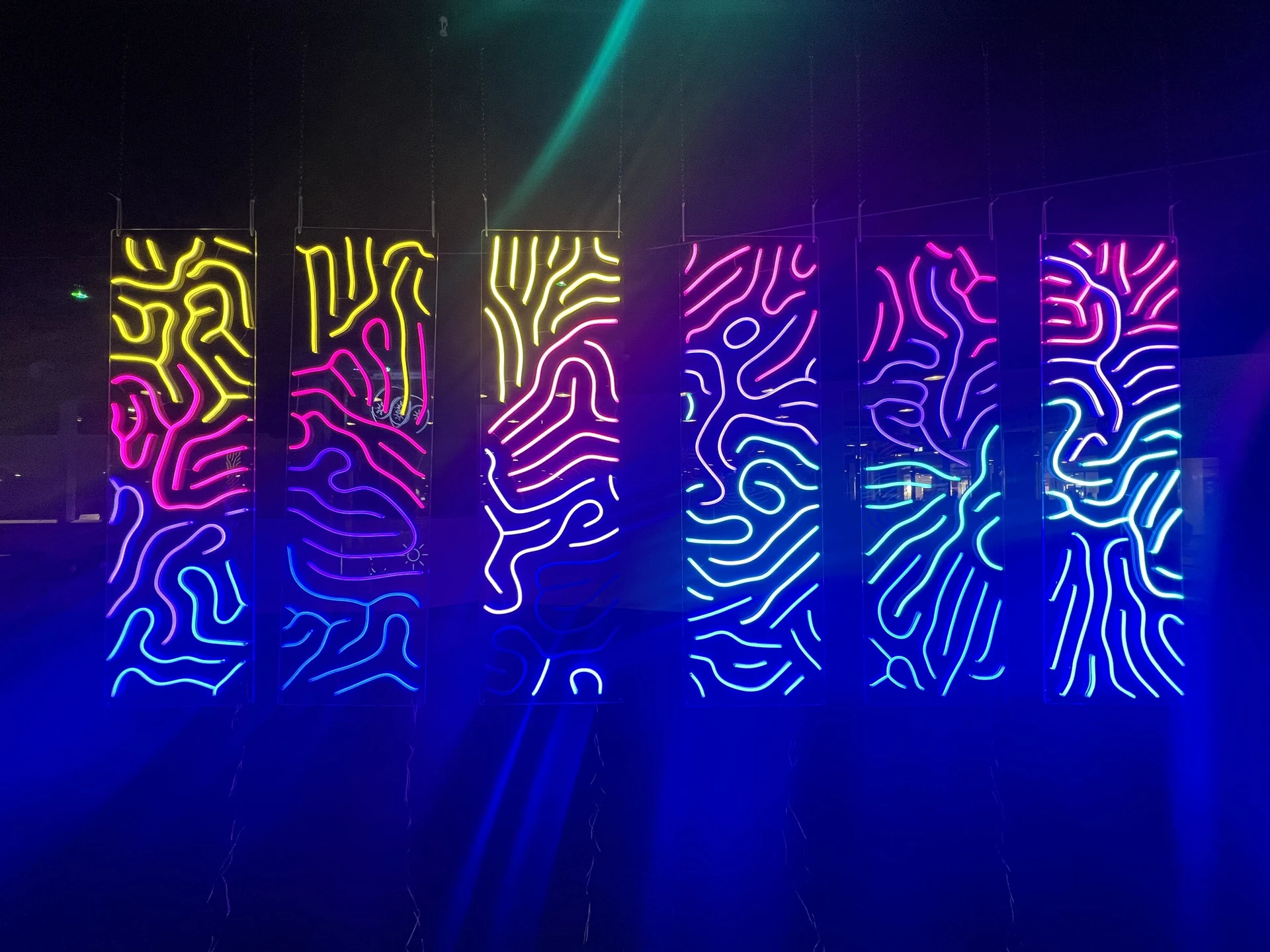 Six hanging neon-like LED signs of winding lines, shading from yellow at the top through pink and violet to blue, in a room washed with blue light.