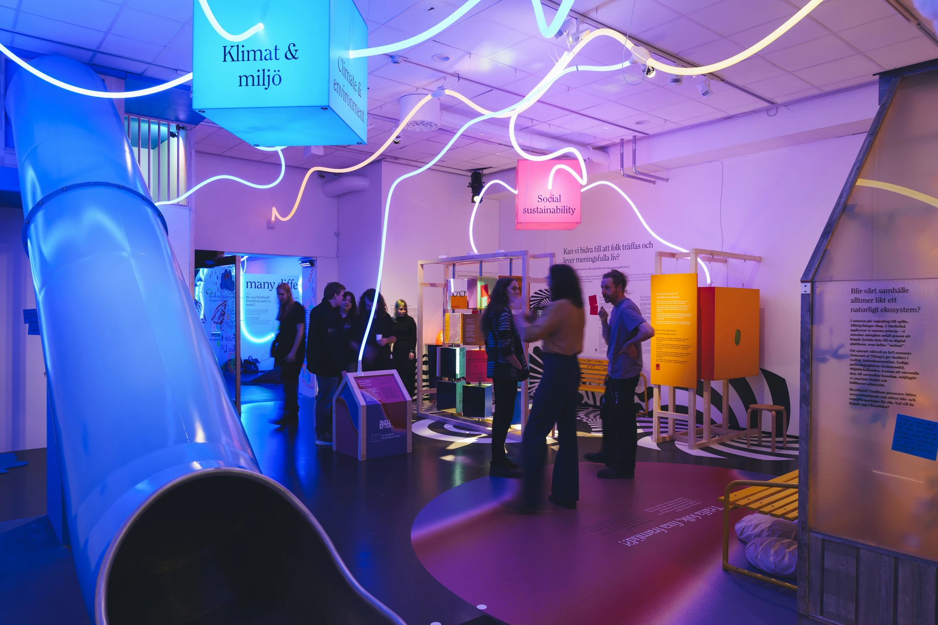 Visitors explore the exhibition beneath winding white lights, with a blue slide and pink, blue and orange displays.